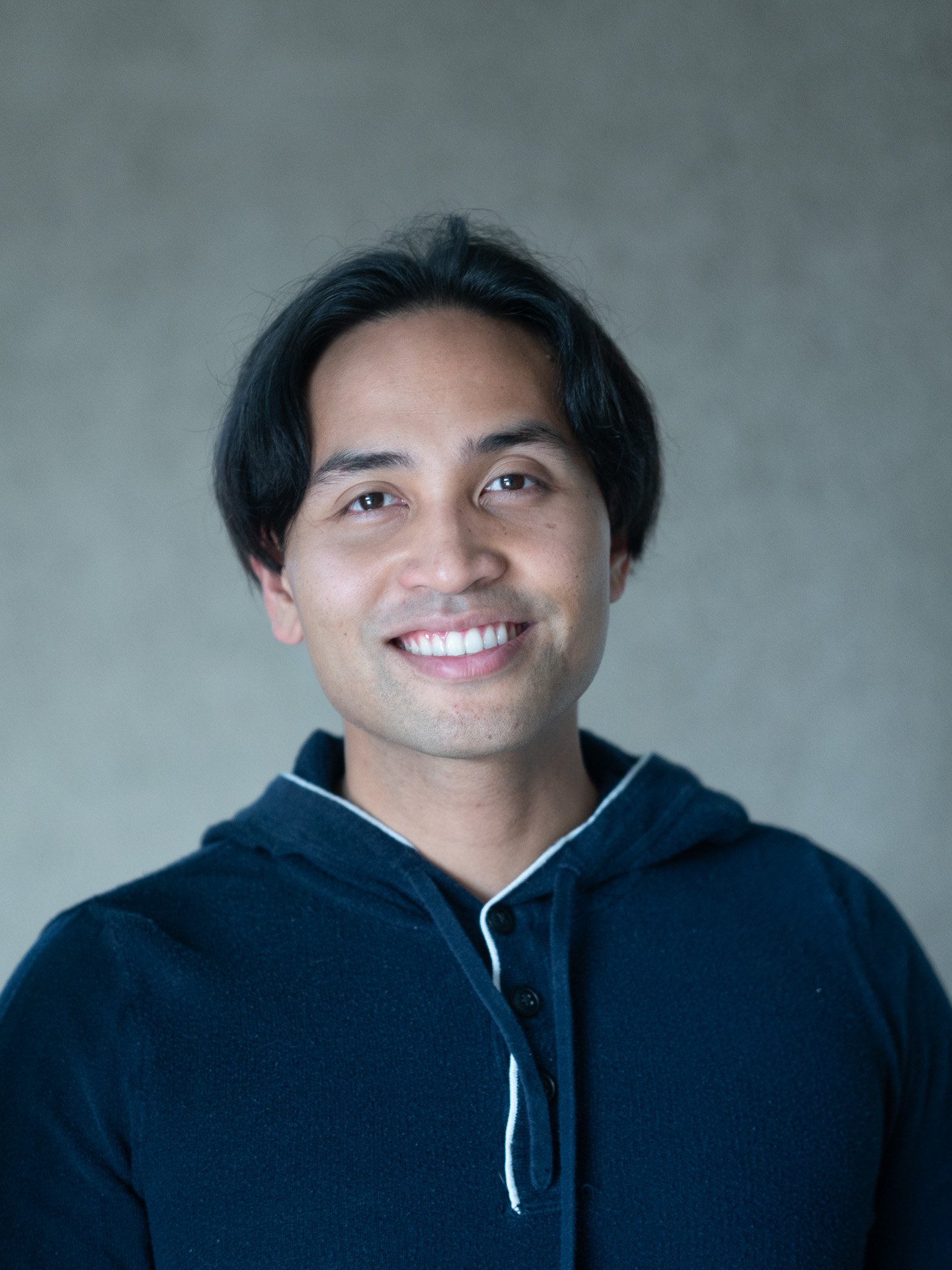 Close-up portrait of a young man with black hair, smiling, wearing a dark blue hoodie with white piping.