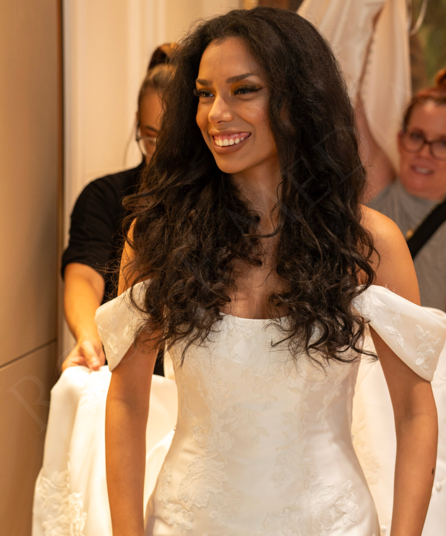 A woman in a white lace dress smiling while trying on wedding gowns with two other women in the background.