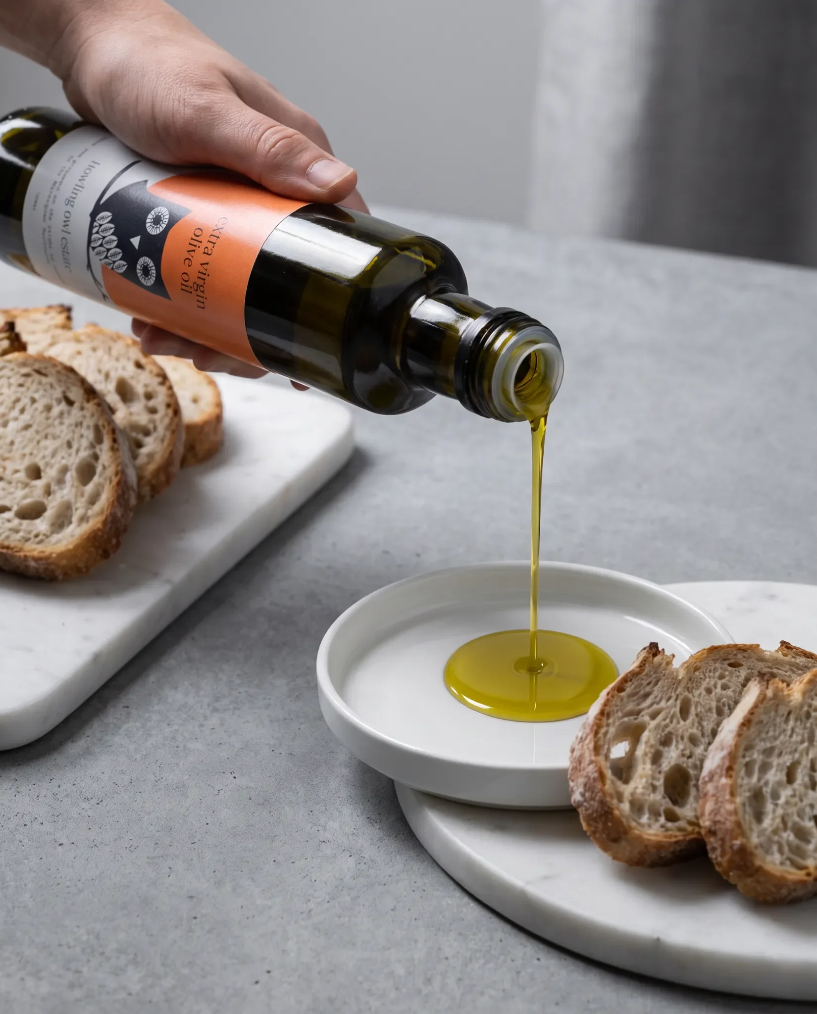 A person pouring olive oil from a bottle into a small white dish, with sliced bread on a white plate in the background.