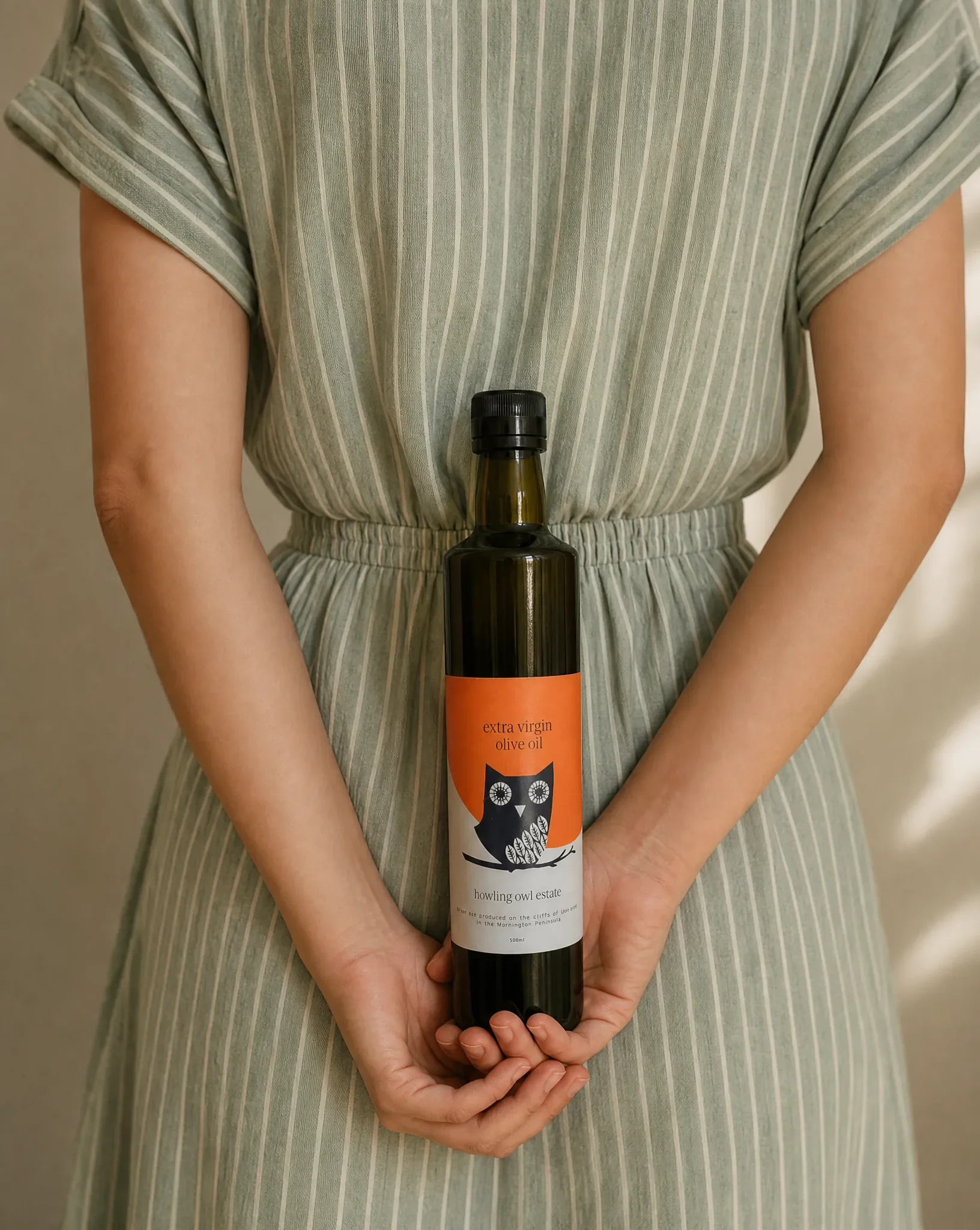 Person in a green and white striped dress holding a bottle of Howling Owl Estate extra virgin olive oil against their waist.
