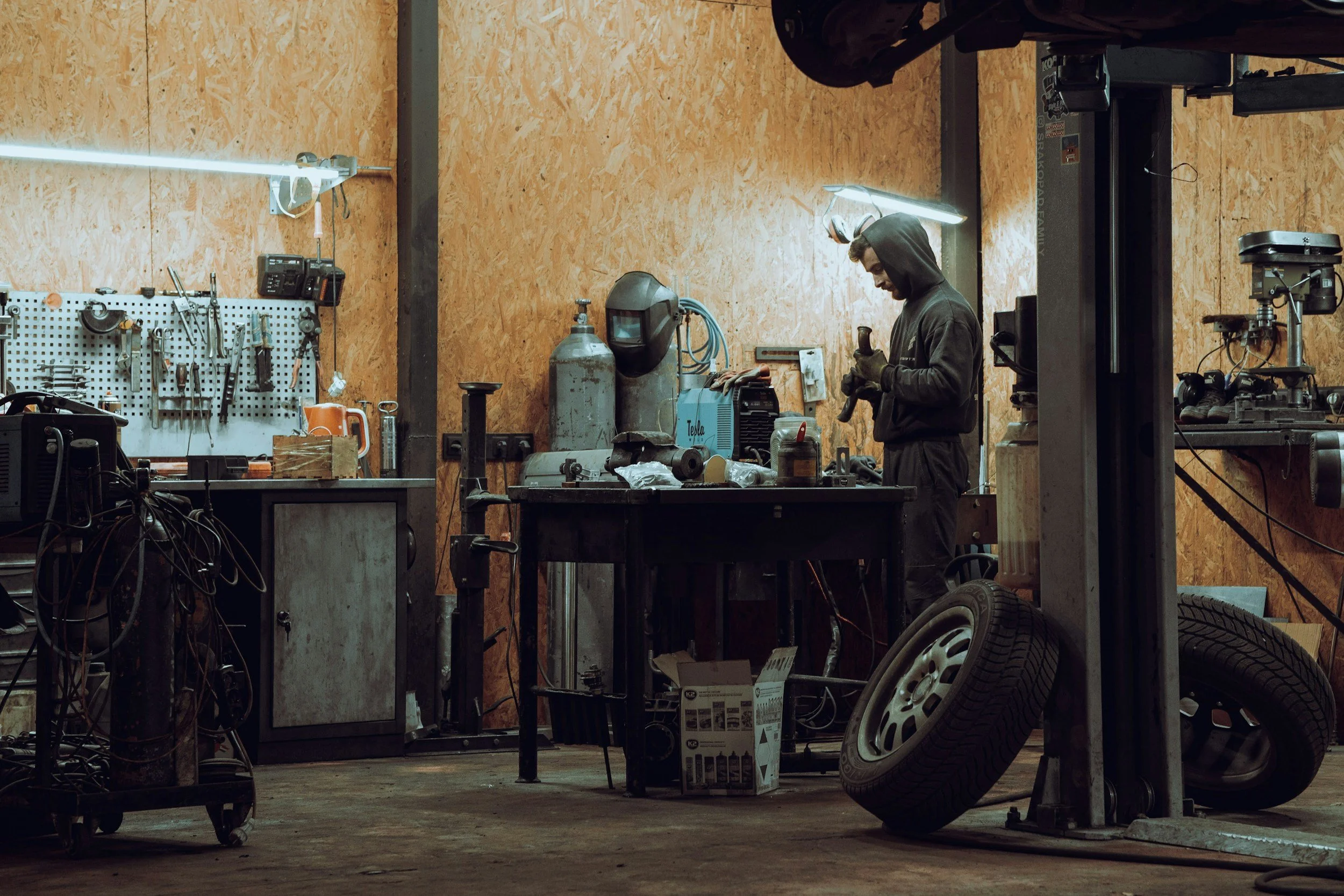 A man working in a workshop with various tools hanging on a pegboard, a gas tank, a welding helmet, and tires on the floor.
