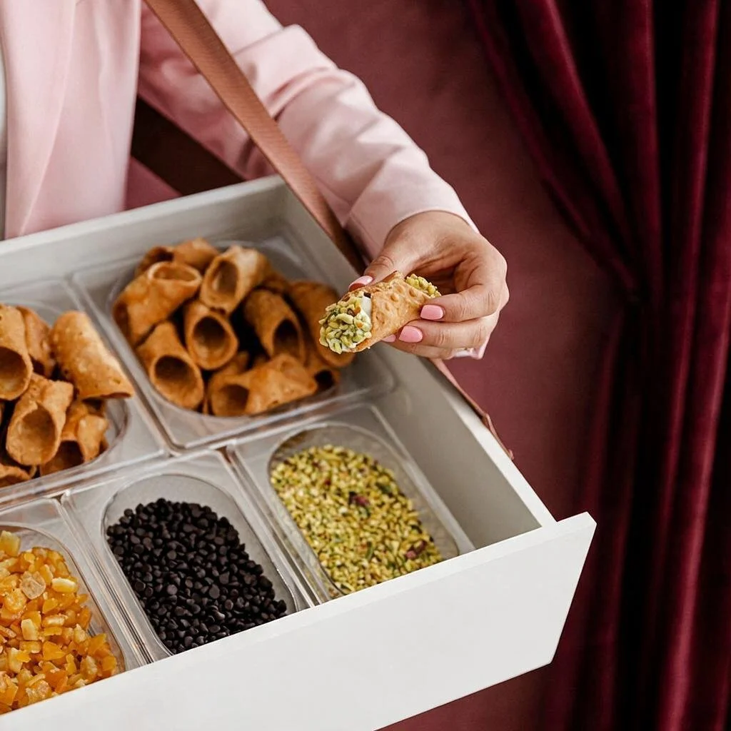 Close-up of a hand holding a pistachio-topped cannoli above a portable dessert cart filled with empty shells and toppings, highlighting the Roaming Cannoli Bar experience at Cannoli Bar Richmond.
