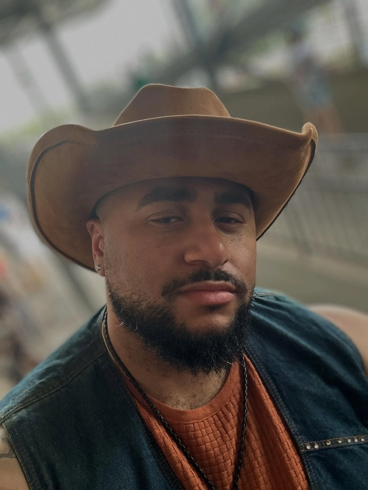 A man with a beard, wearing a cowboy hat, a black leather jacket, an orange shirt, and a necklace.
