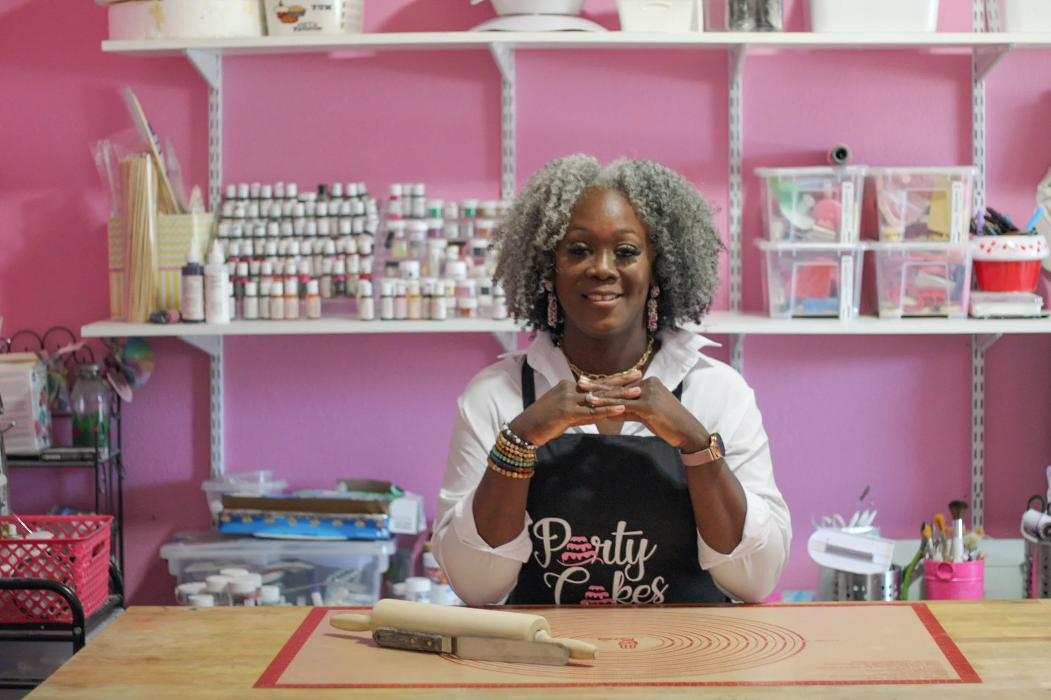 A smiling African American woman with gray curly hair sitting at a wooden table in a pink-themed baking or cooking space. She is wearing a white shirt and a black apron with the word "Party" on it. On the table in front of her is a rolling pin, and the background features shelves filled with small containers of colorful sprinkles and baking supplies.