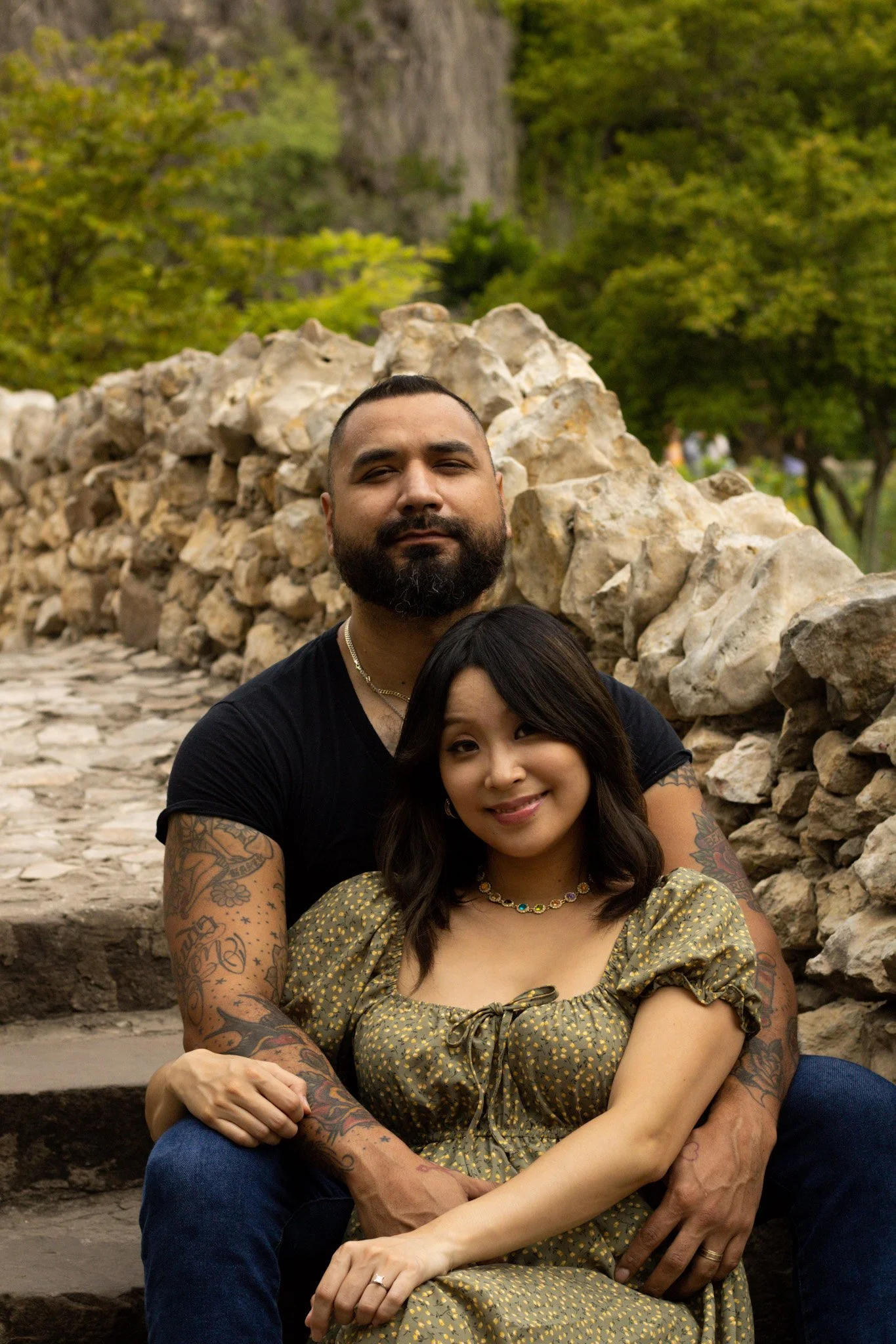A couple sitting outdoors in front of a stone wall and green trees, smiling at the camera.
