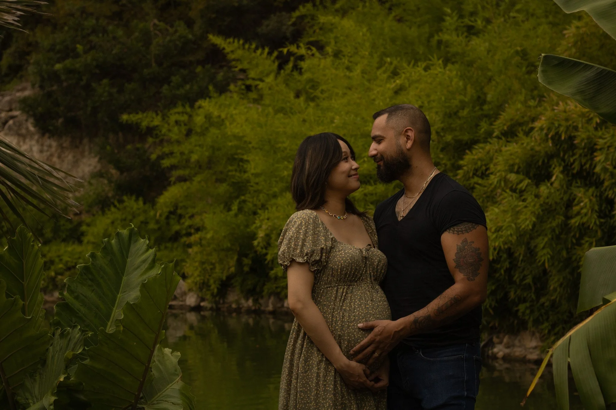 A couple standing close together outdoors, facing each other and smiling, surrounded by lush green vegetation.