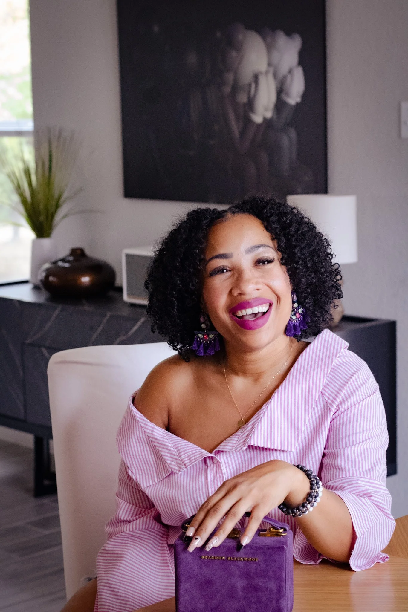 Smiling woman with curly hair, wearing purple tassel earrings and a pink and white striped off-shoulder top, sitting at a table with a purple book titled 'Brandon Blackwood' in a modern living room with artwork on the wall and a vase with green plants in the background.