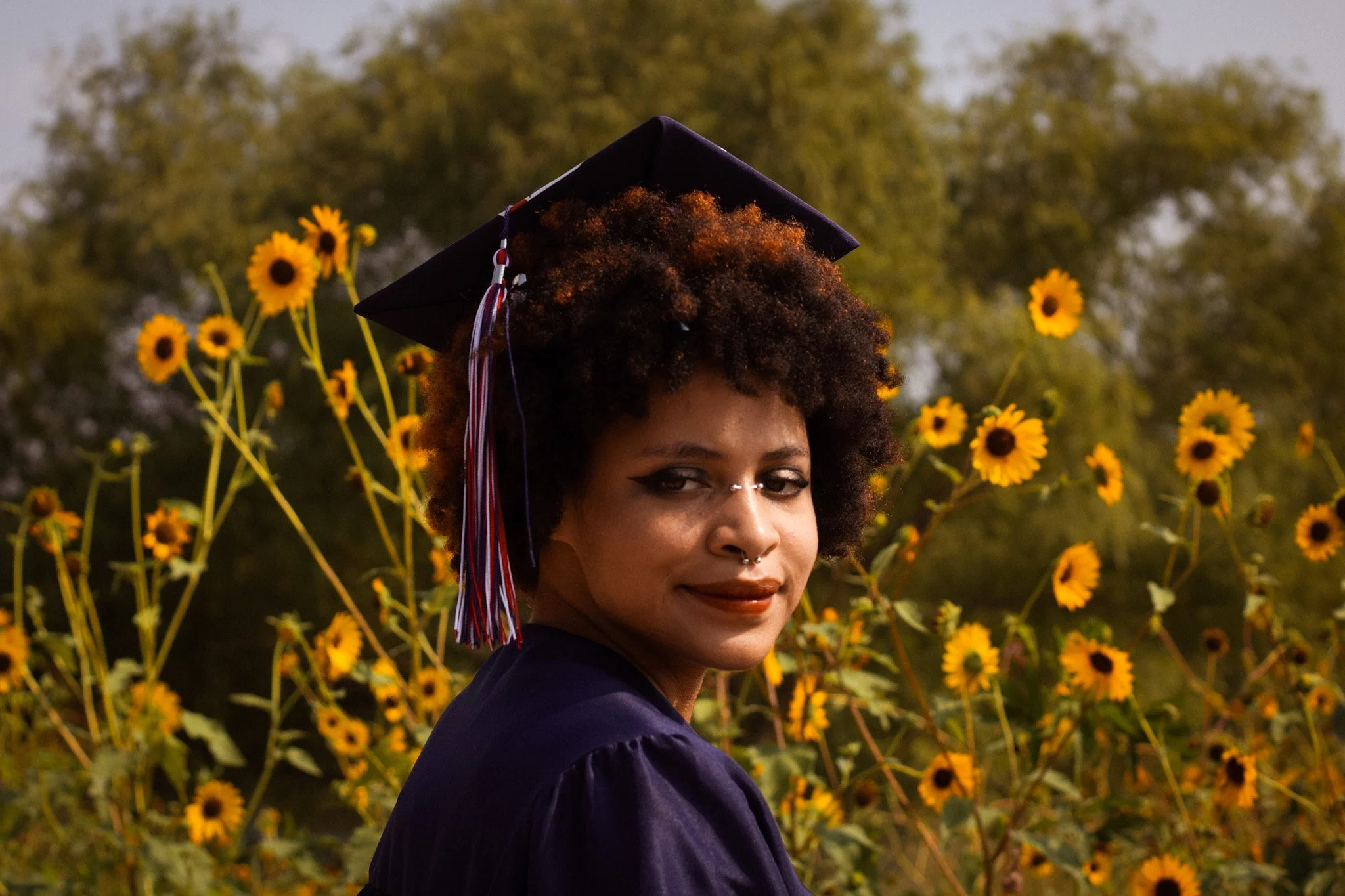 A woman with curly hair and multiple piercings, wearing a graduation cap and gown, standing in front of a field of yellow flowers during sunset.