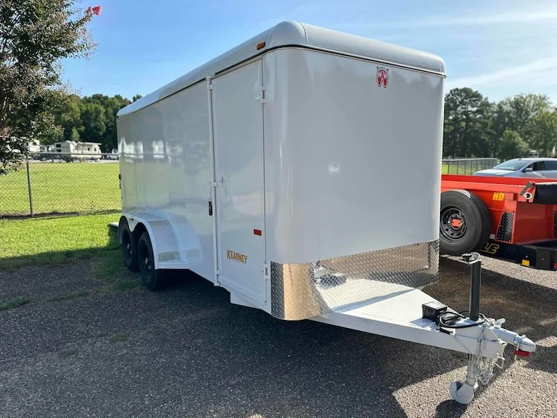 White enclosed cargo trailer with dual axles parked on gravel, with a red vehicle partially visible next to it and trees and grass in the background.