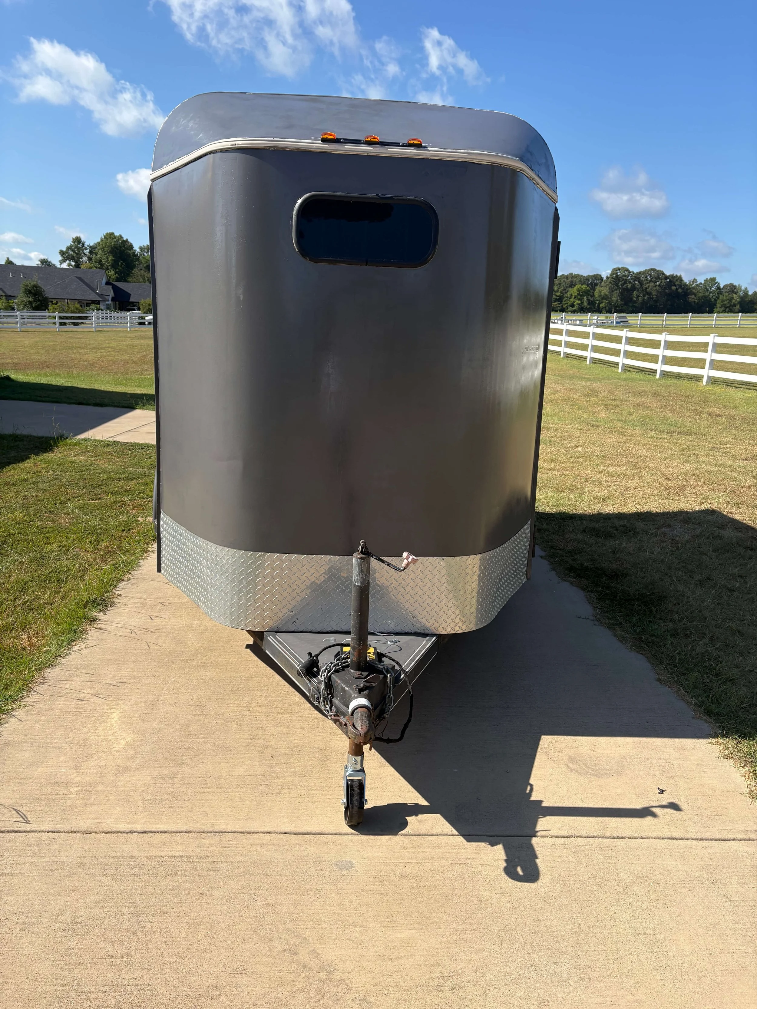 A black and silver enclosed trailer attached to a hitch on a concrete driveway, with a grassy area, white fence, trees, houses, and a blue sky with scattered clouds in the background.
