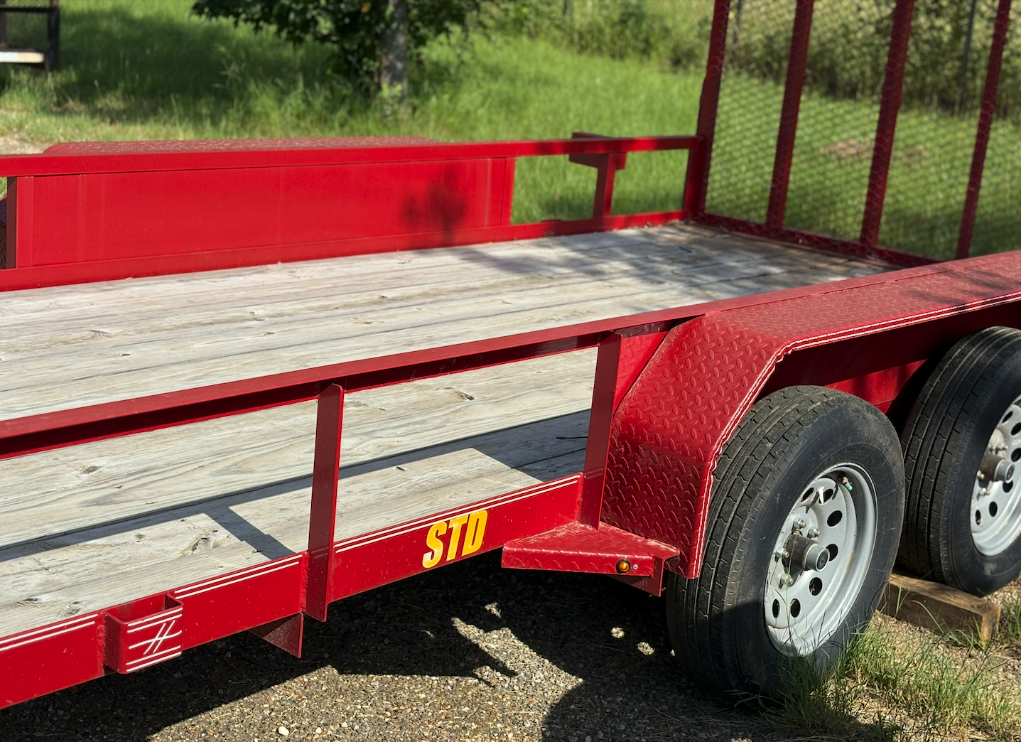 Red flatbed trailer with wooden deck and dual wheels parked on a gravel surface, with green grass and trees in the background.