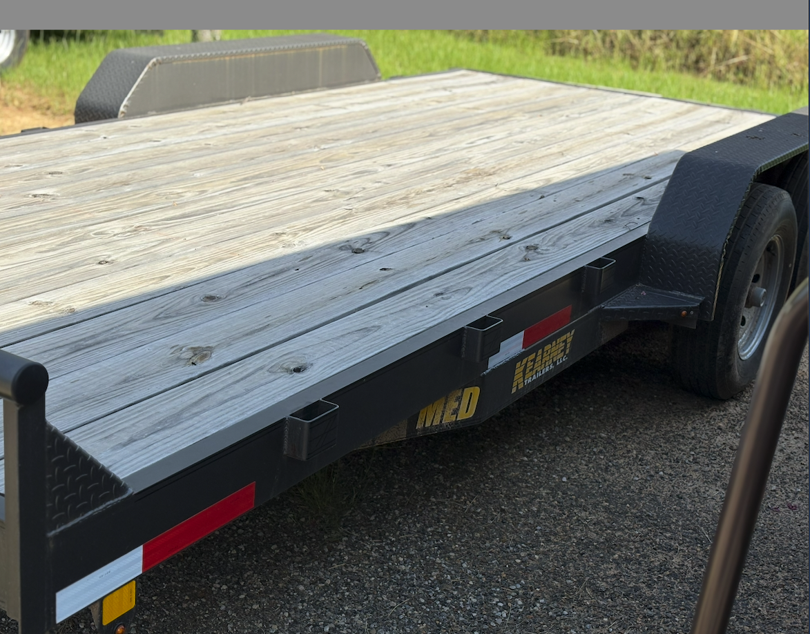 A flatbed trailer with a wooden deck and black fenders parked on a gravel surface with grass in the background.