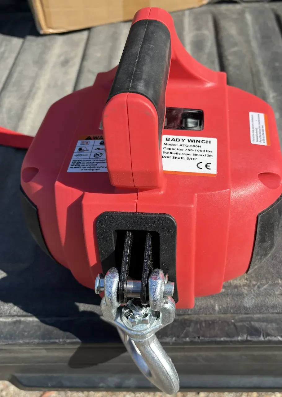Red cordless baby winch with black handle, warning labels, and metal hook, placed on a wooden surface.