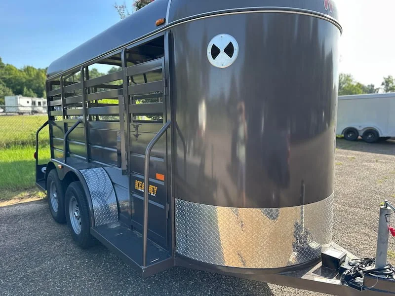 Black livestock trailer with open sides, metal slats, rounded front, and a cartoon skull logo on the front. Trailers seen in the background, parked on gravel ground.
