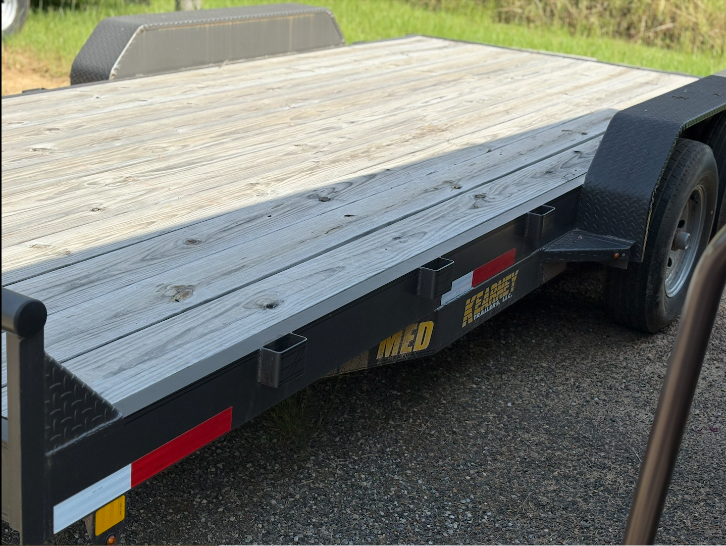 A black flatbed trailer with a wooden deck parked on gravel, with grass and trees in the background.