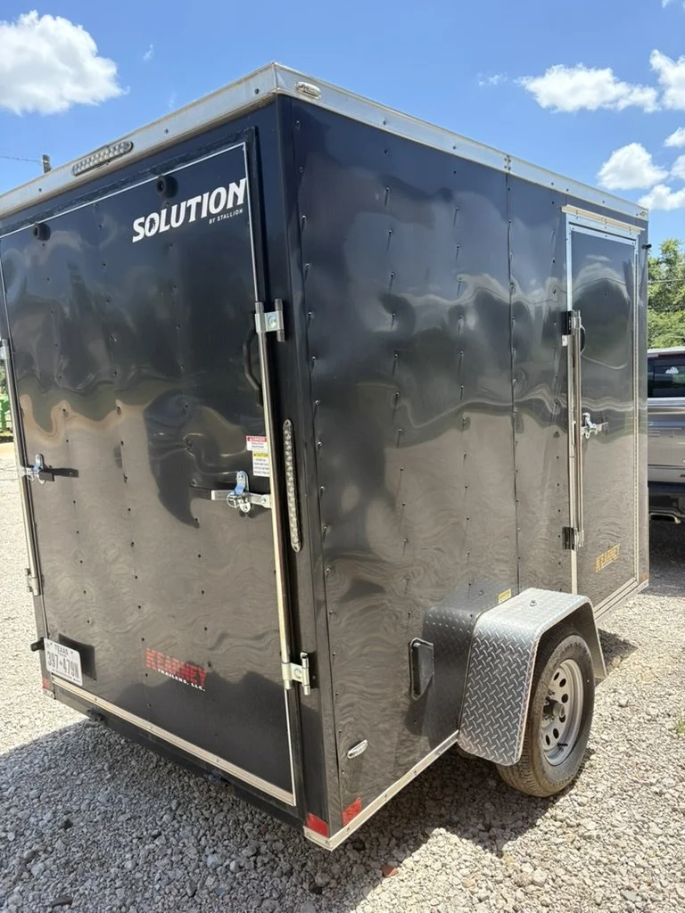 Black enclosed trailer with a single axle, parked on gravel under a partly cloudy sky.