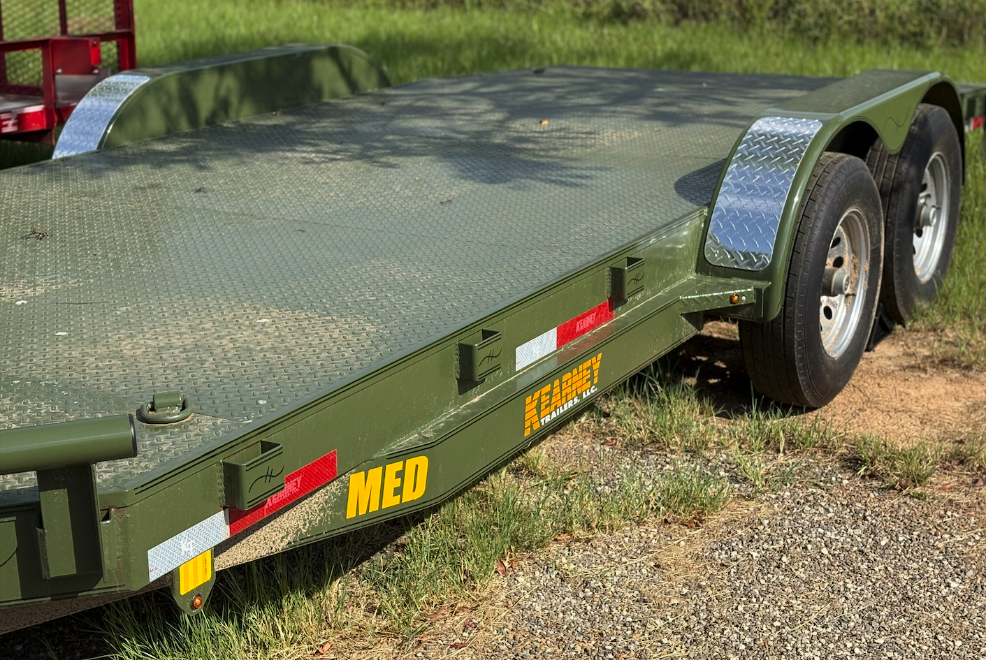 A green flatbed trailer with dual axles parked on grassy and dirt ground, with equipment and grass in the background.