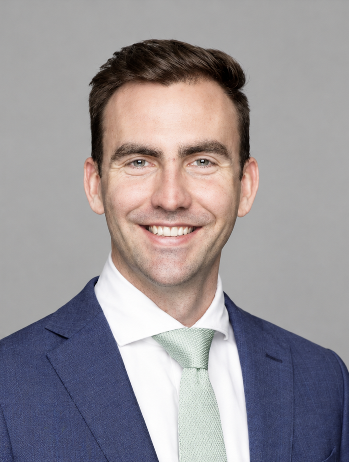 A professional headshot of a smiling man in a blue suit, white shirt, and light green tie against a gray background.