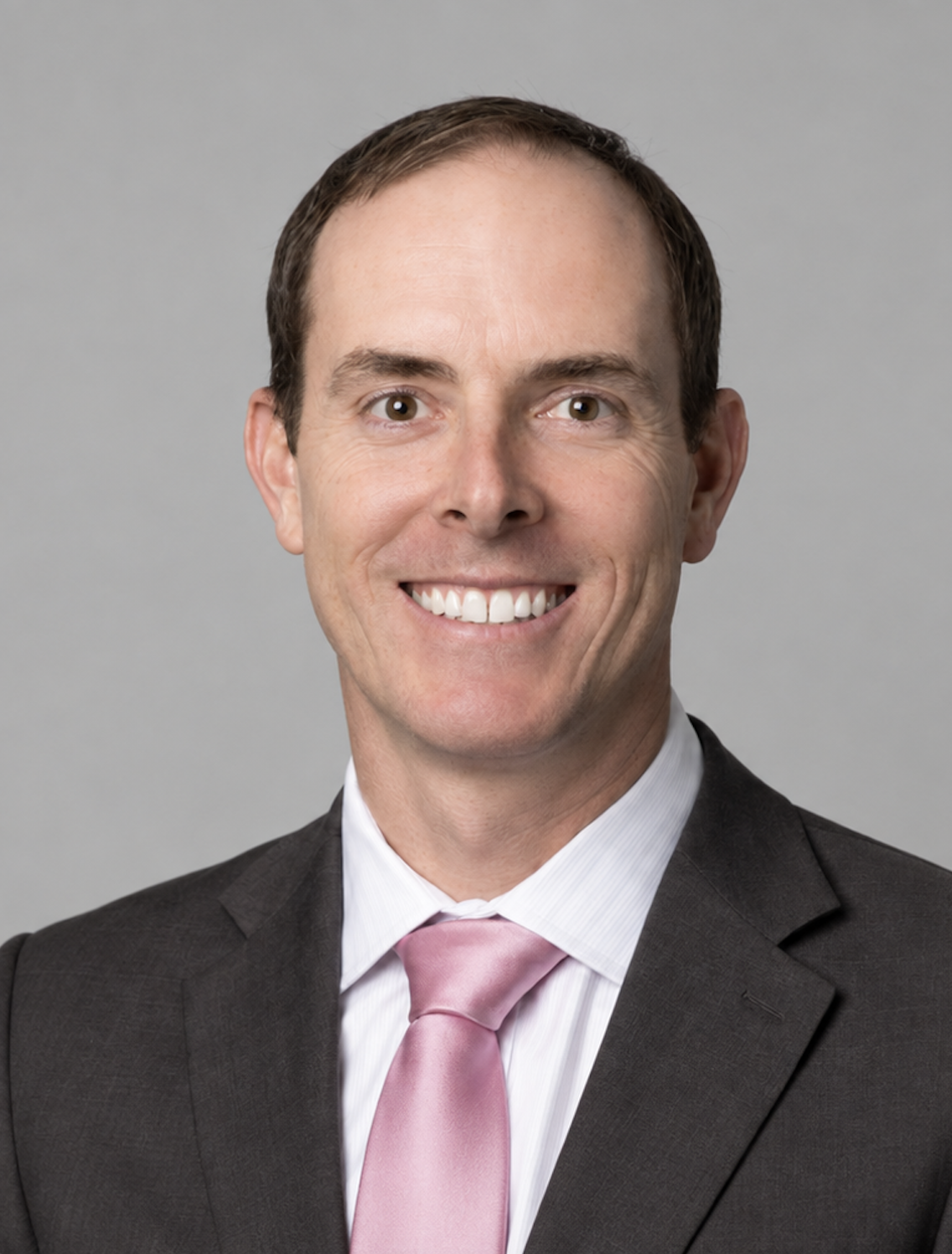 Headshot of a man in a suit with a pink tie, smiling against a gray background.