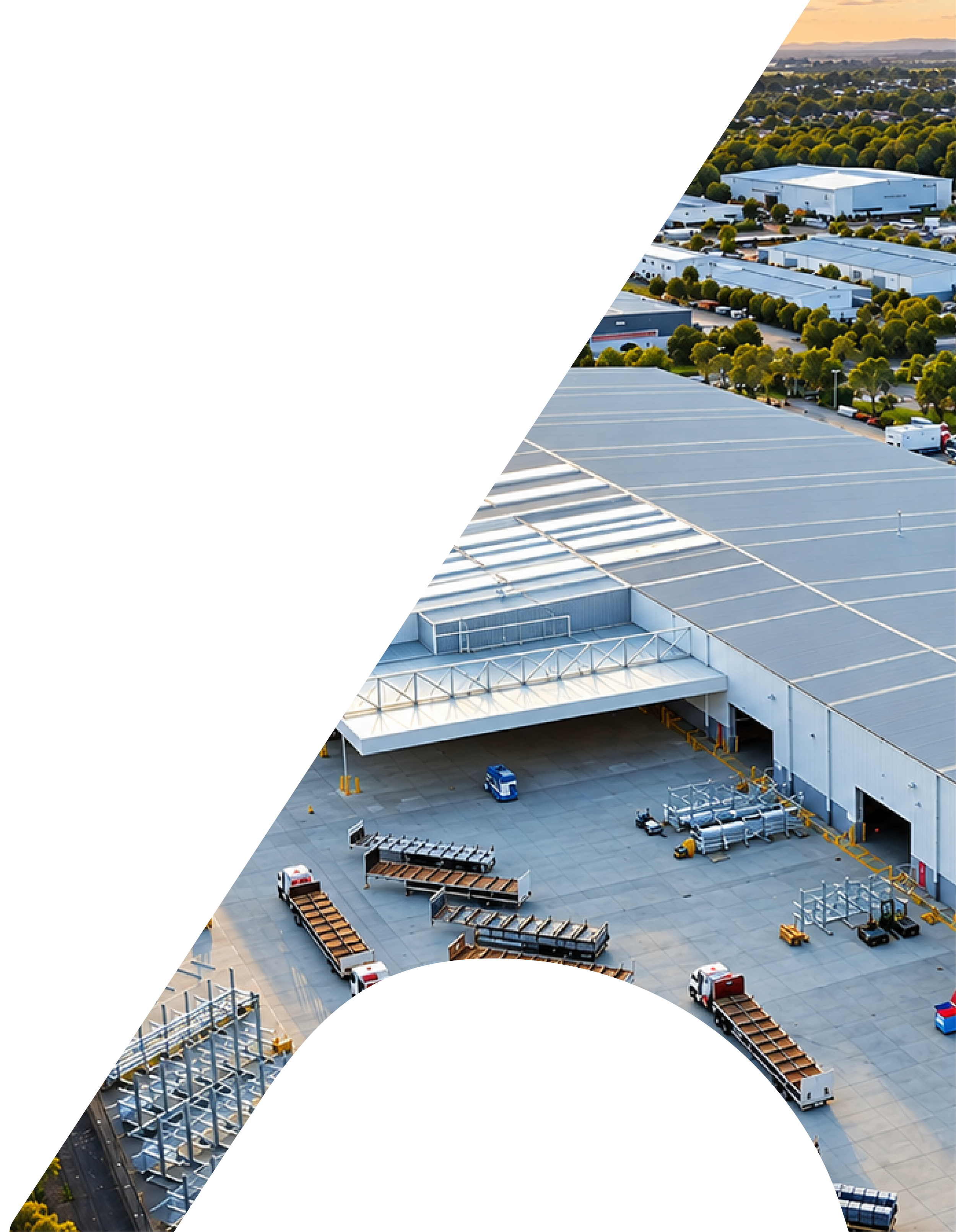 Aerial view of a large logistics warehouse with trucks and cargo containers, some parked outside and others on internal loading docks, surrounded by green trees and a distant mountain range at sunset.