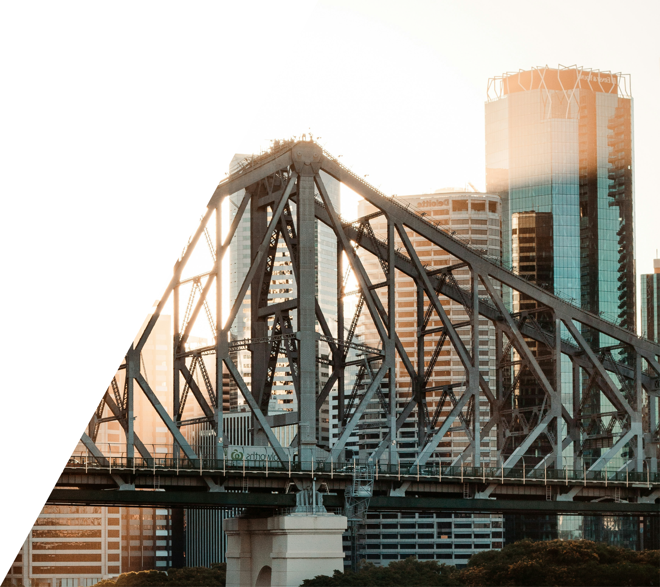 A city skyline during sunset featuring a large steel bridge with multiple support beams in the foreground and modern glass high-rise buildings in the background.