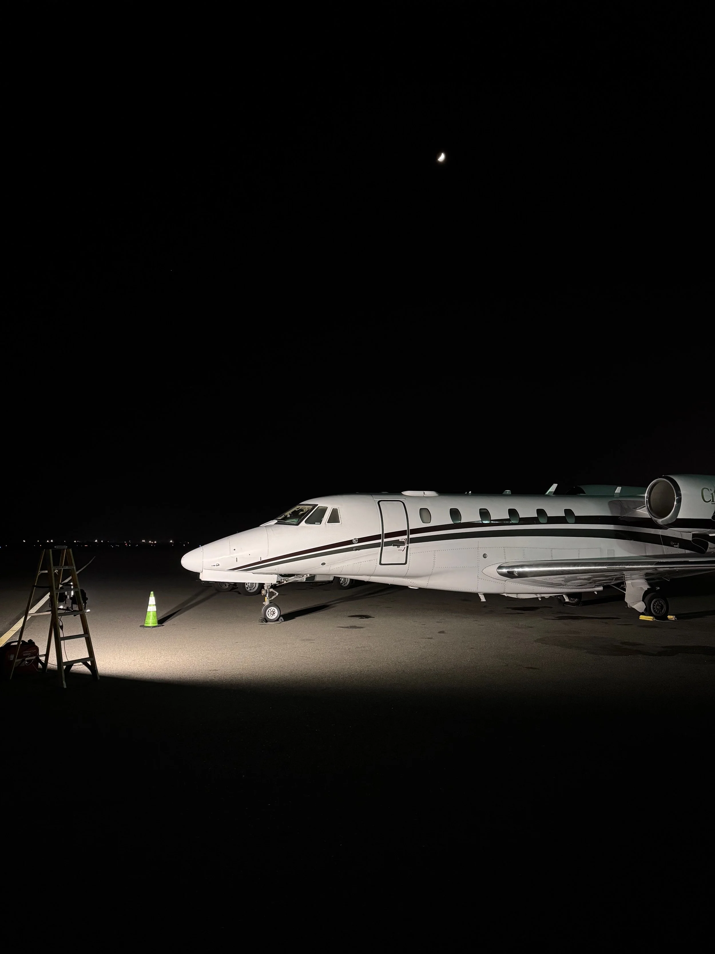 A private jet aircraft parked on the tarmac at night, illuminated by ground lights, with airport equipment and cones nearby.