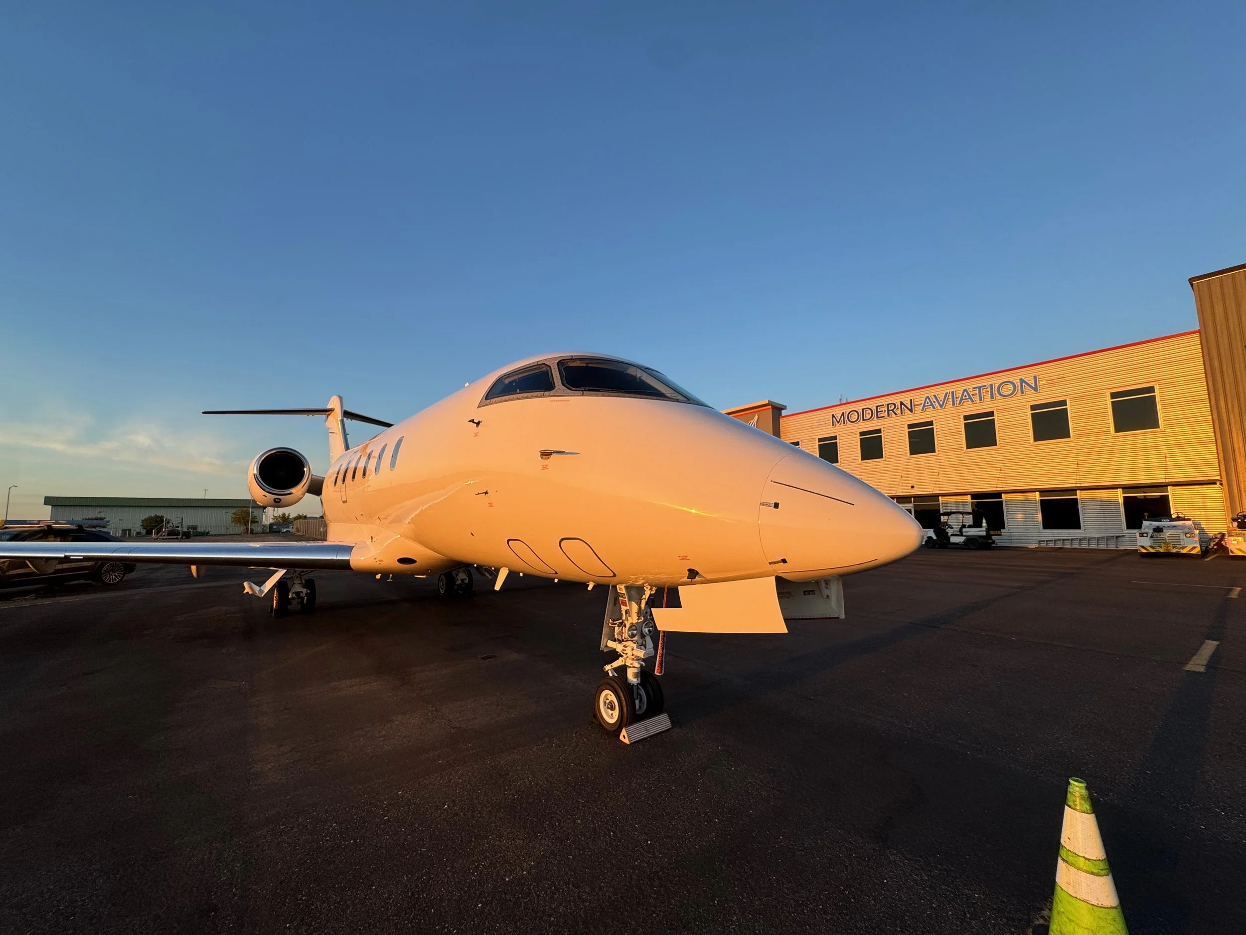 Private jet parked at an airport during sunset, with a building labeled 'Modern Aviation' in the background and traffic cones in the foreground.