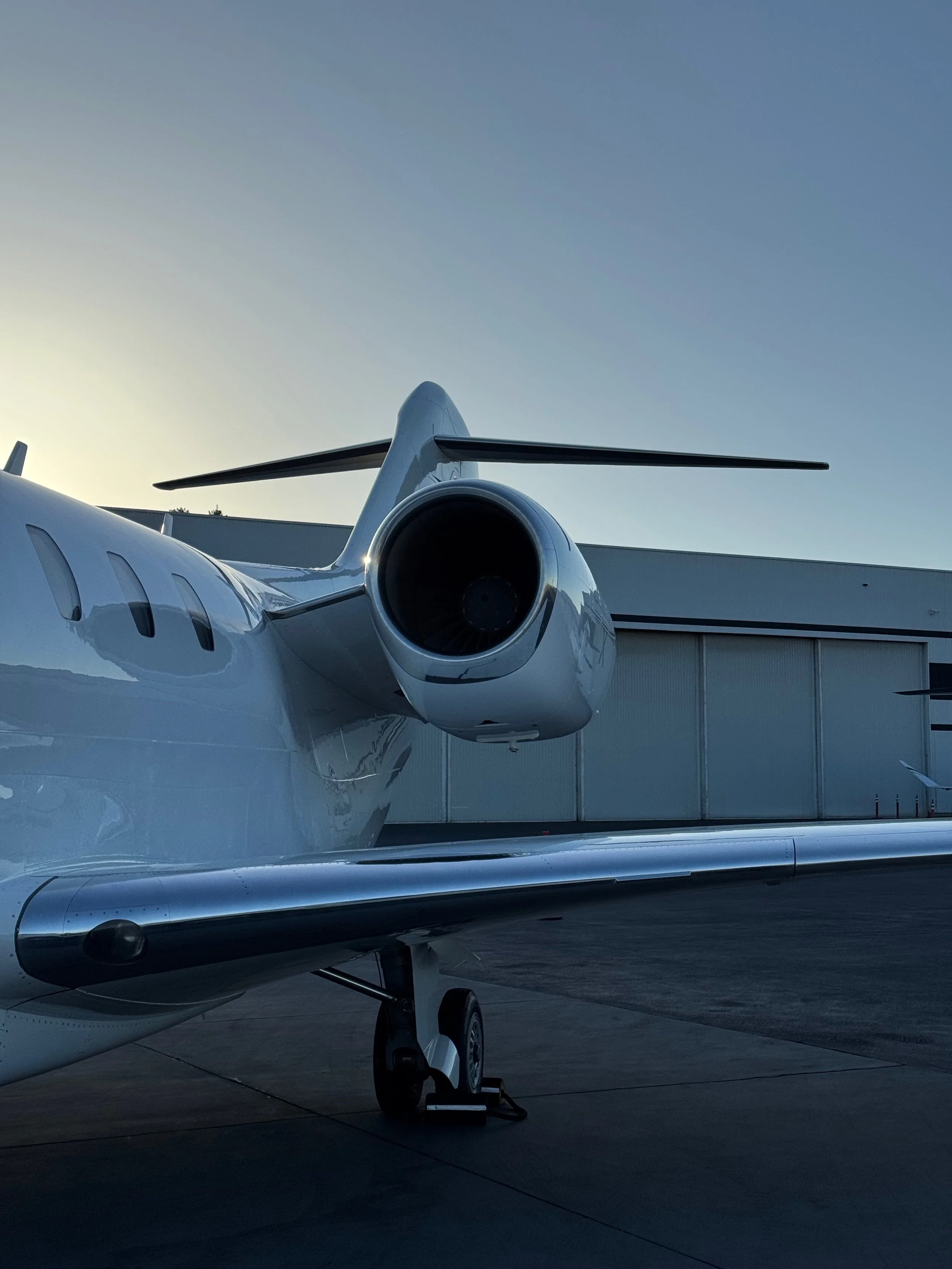 Rear view of a white private jet parked on the tarmac during sunset or sunrise, with a clear sky in the background.