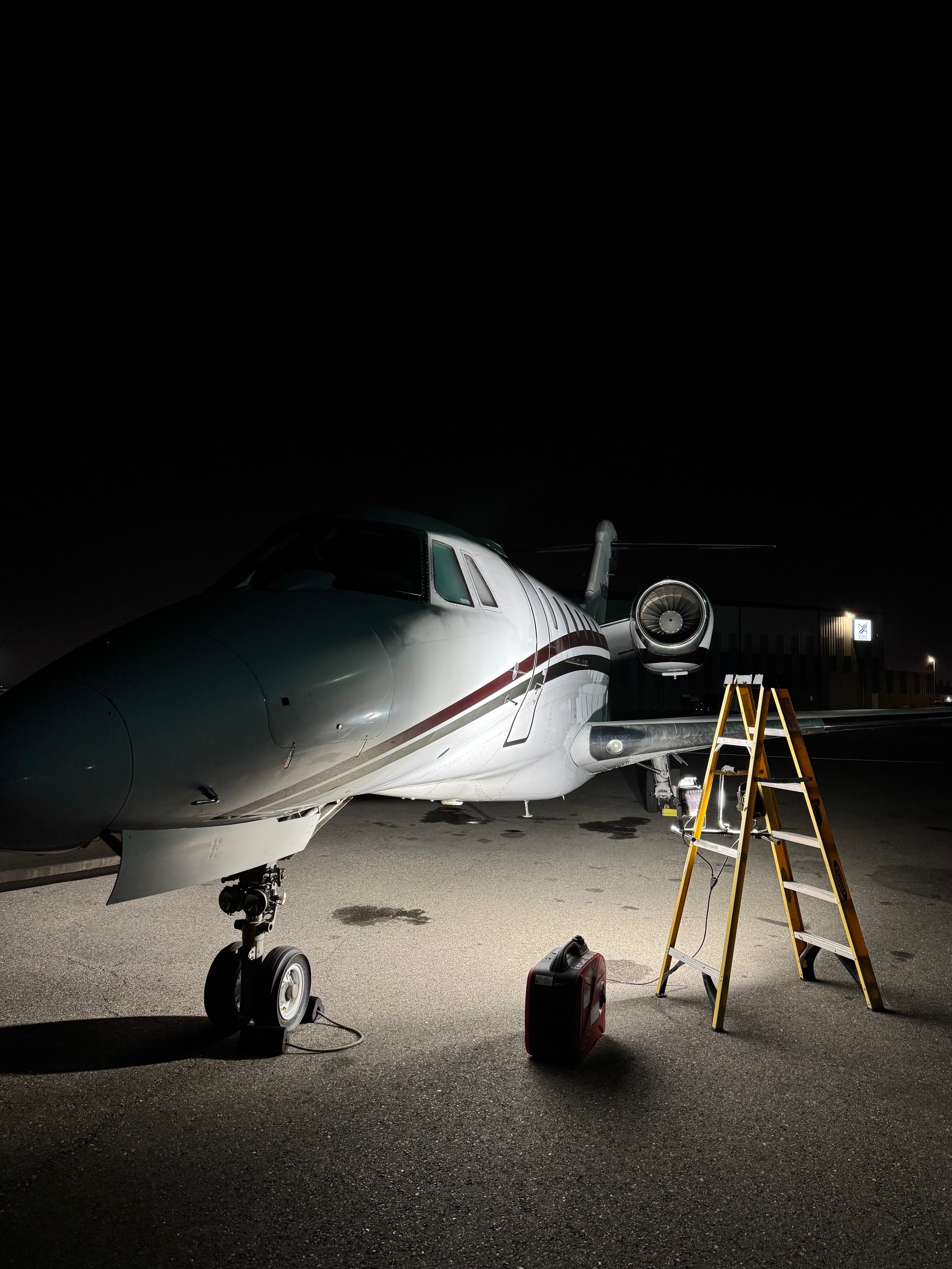 A private jet parked on the tarmac at night with maintenance equipment and a yellow ladder nearby.