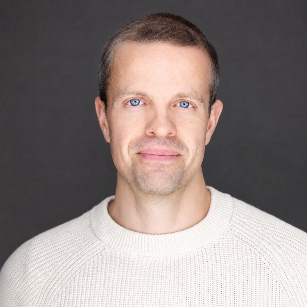 Portrait of veroFresh's founder, Mark Hogan, with short brown hair and bright blue eyes, wearing a cream-colored sweater, smiling lightly against a dark gray background.
