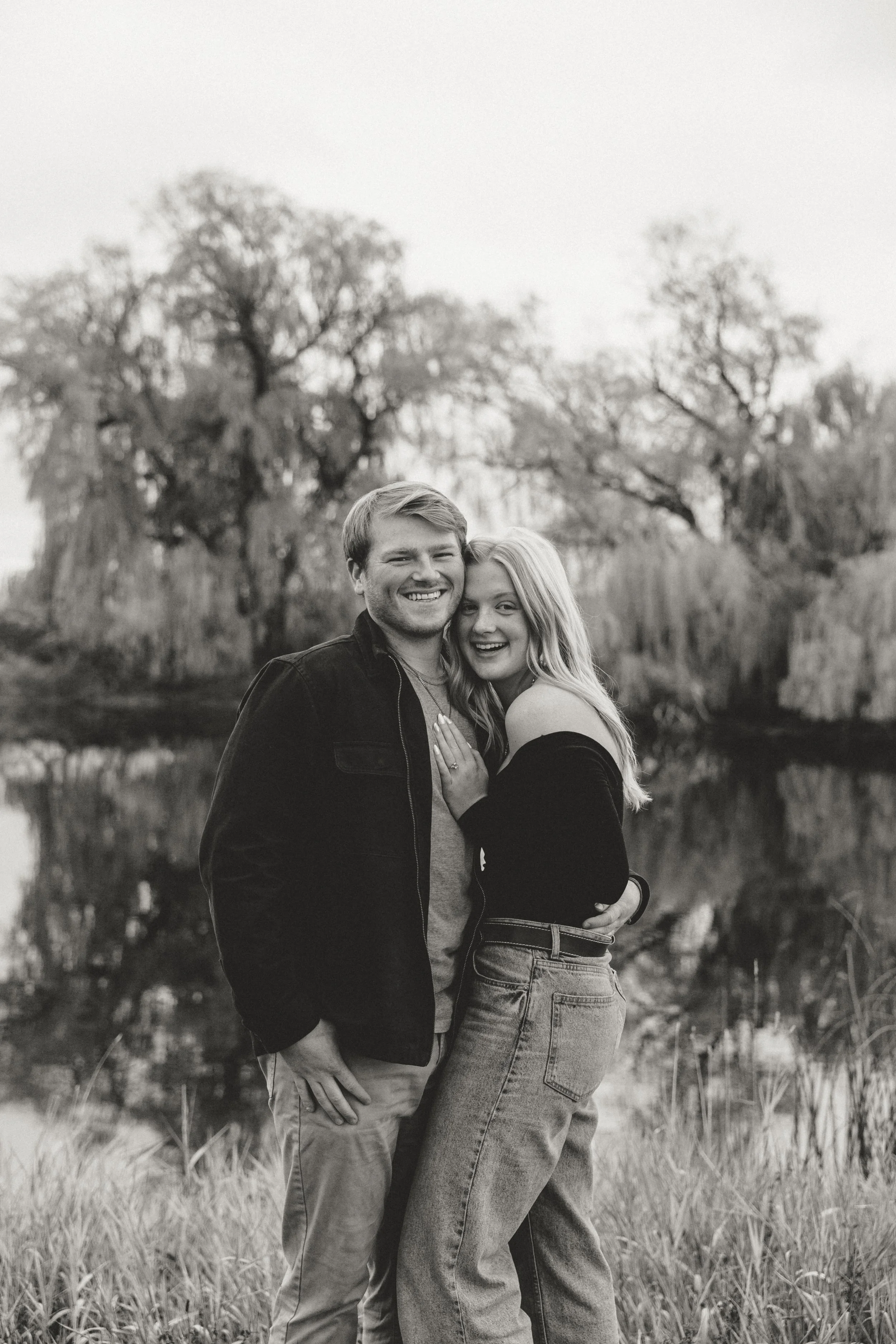 A black and white photo of a smiling man and woman embracing outdoors by a pond with trees in the background.