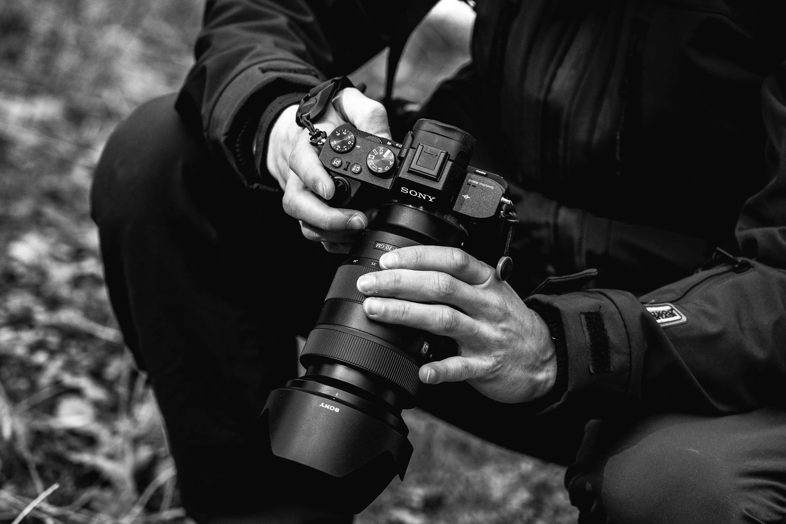 Close-up of a person wearing dark outdoor clothing, holding a Sony mirrorless camera with a large lens in their hands. The background shows a blurred outdoor setting.