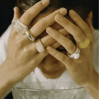Person with hands covering face, wearing large rings, sitting at a table with a glass bowl in front.