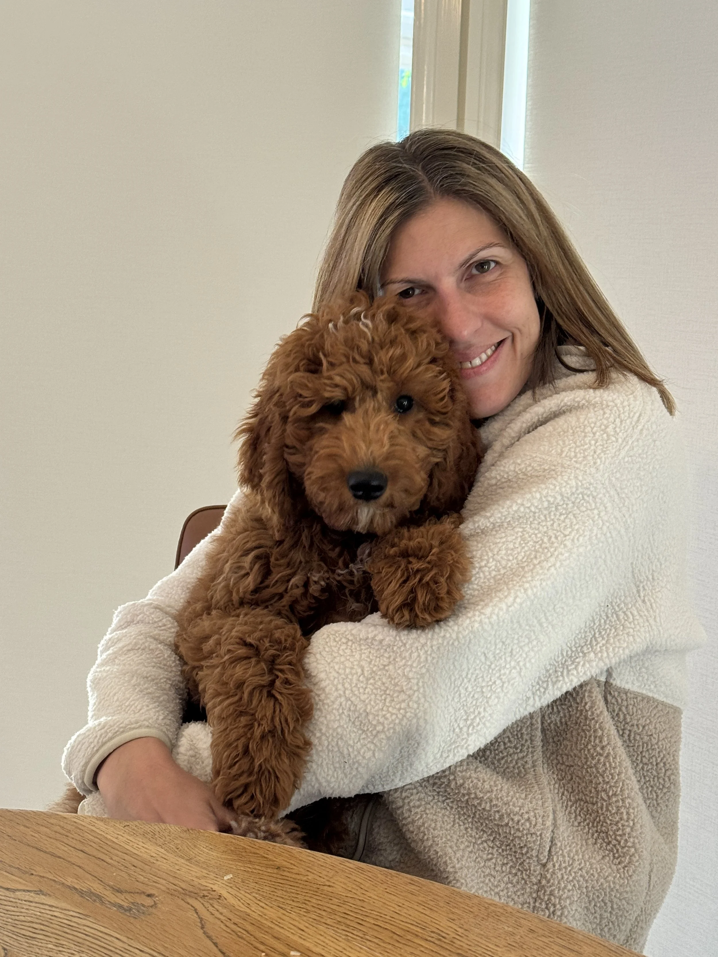 A woman smiling while holding a fluffy, brown puppy close to her face indoors near a window with white walls and a wooden table in the foreground.