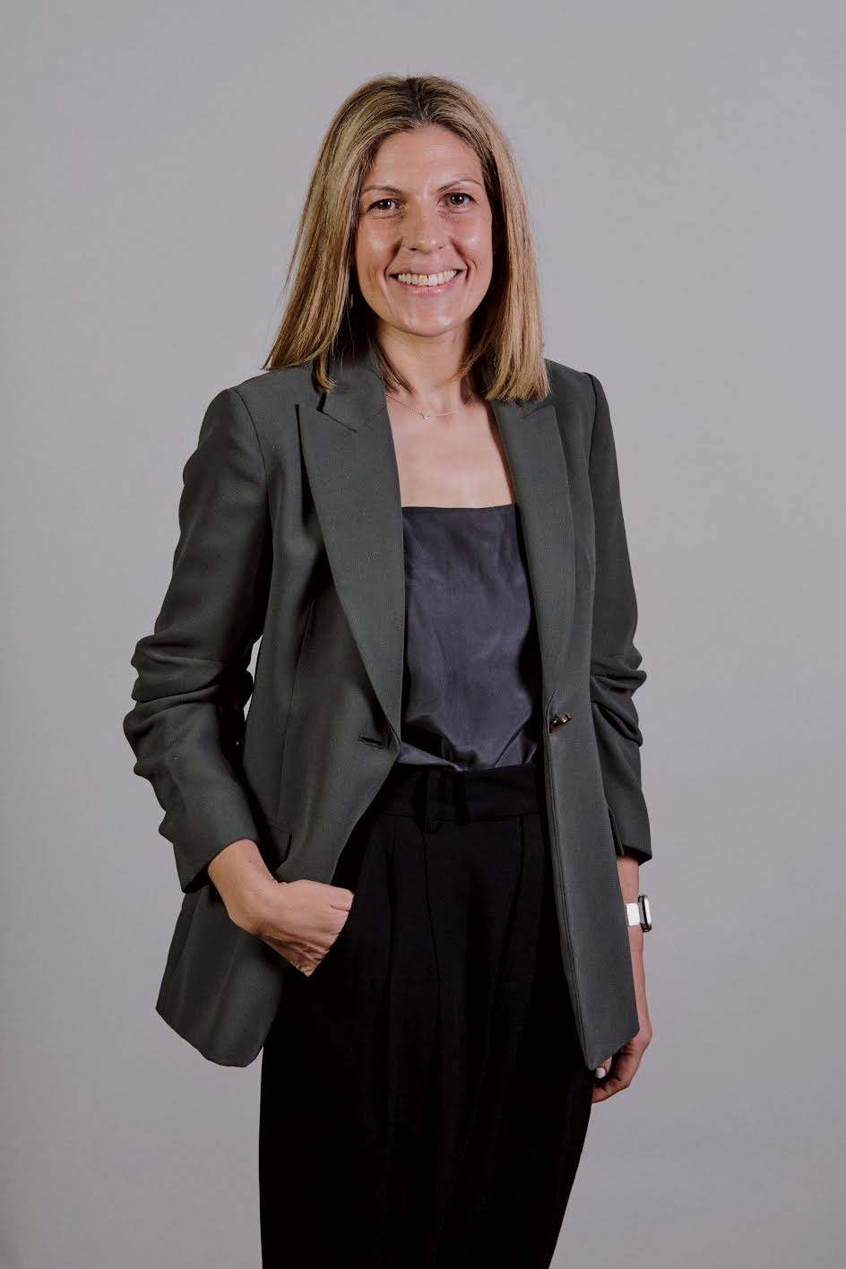 A smiling woman with shoulder-length blonde hair, wearing a dark blazer, black blouse, and black trousers, standing against a plain grey background.