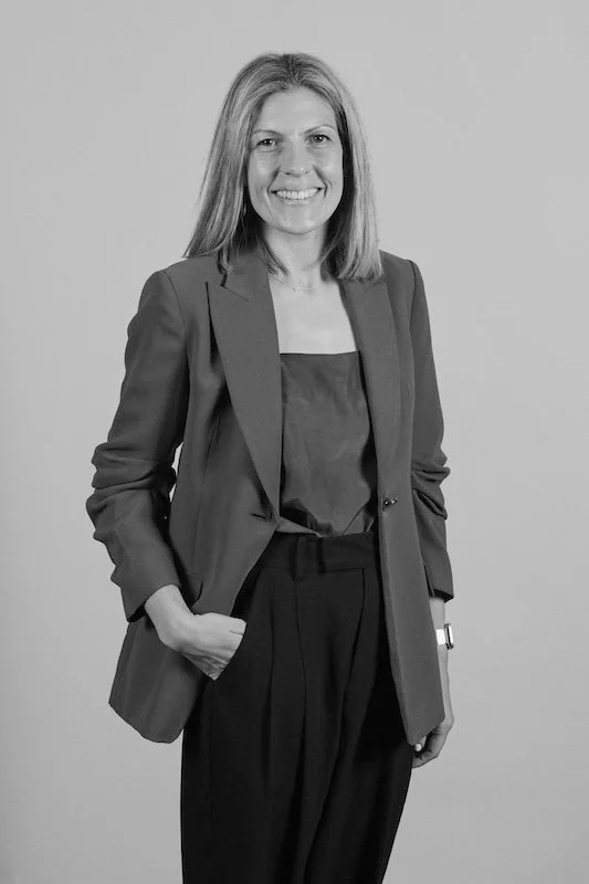 Black and white photo of a woman in business attire, smiling, with one hand in her pocket, standing against a plain background.