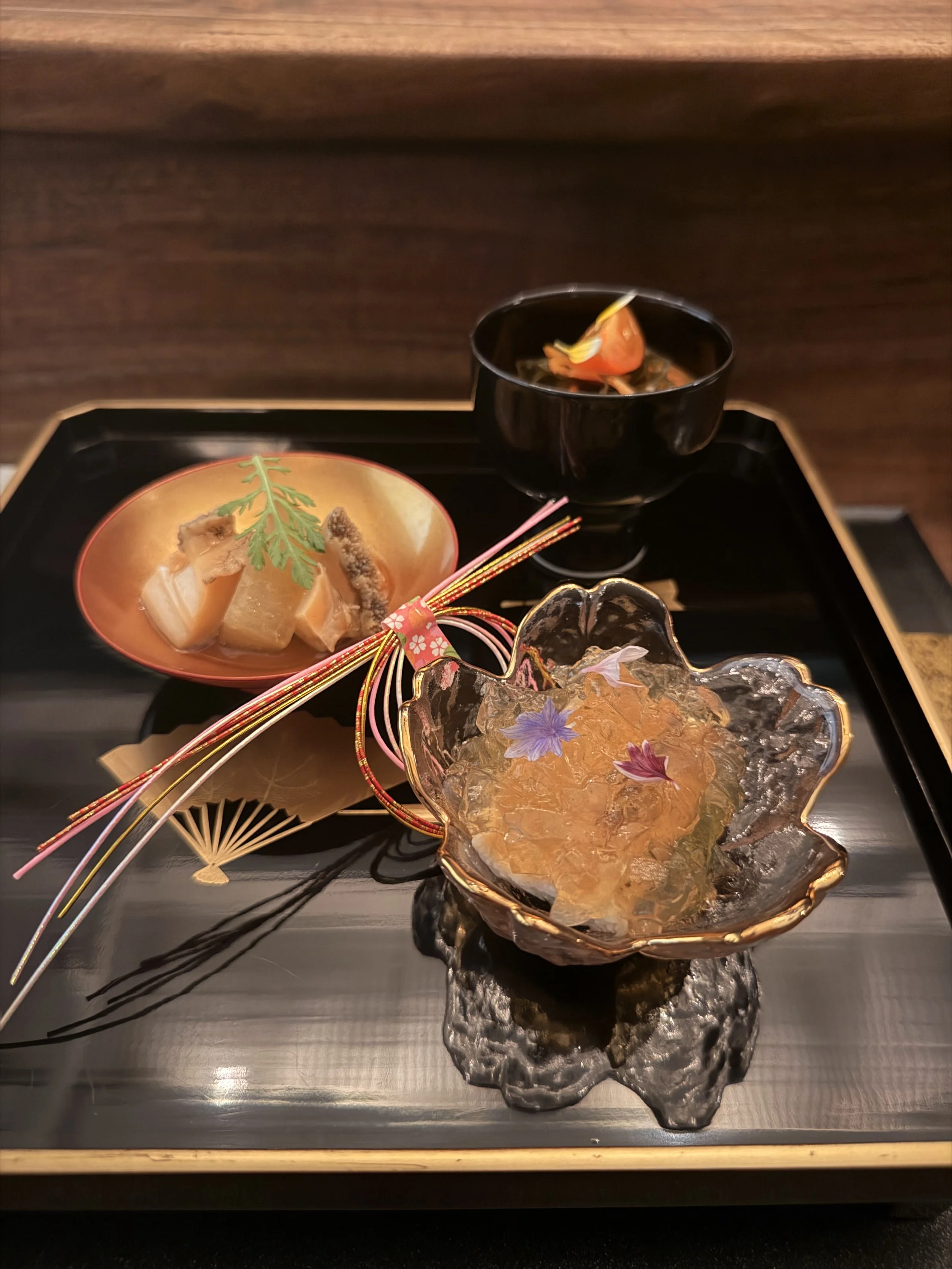 A traditional Japanese meal featuring a black bowl with a seafood dish and a decorative glass dish filled with dashi jelly on a black tray.