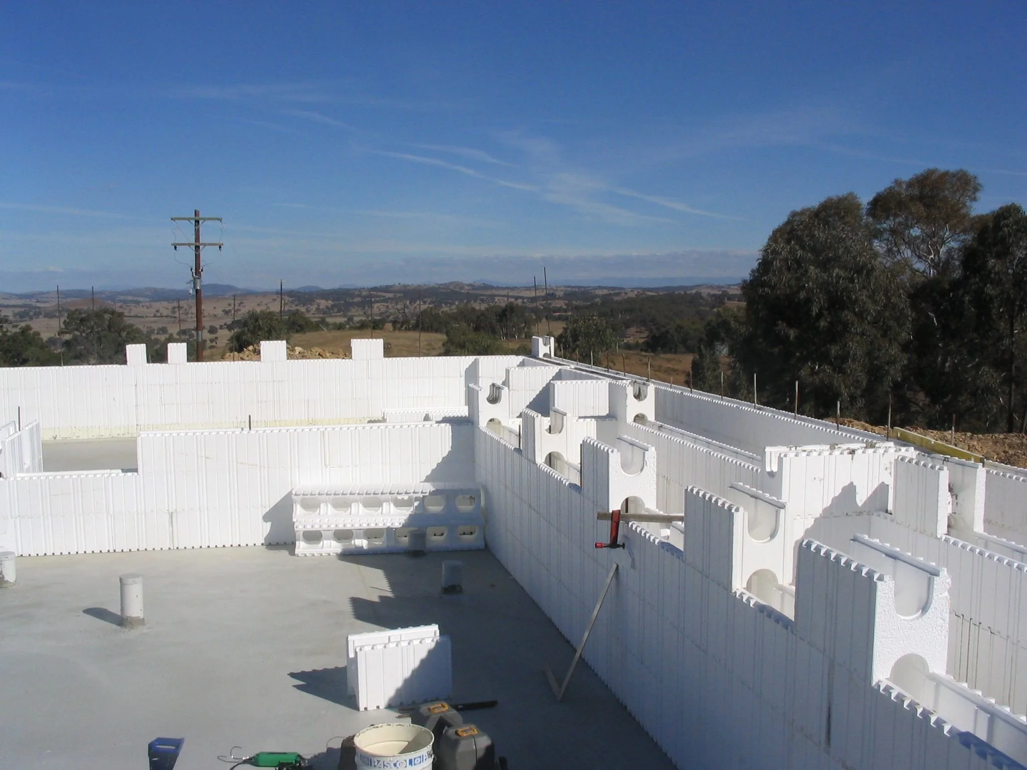 White plastic fencing being assembled on a concrete surface with tools laying on the ground, set against a rural landscape with trees, rolling hills, and a blue sky.