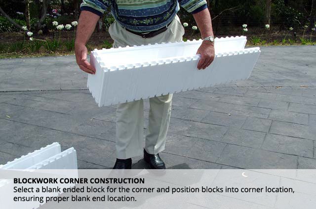 A person holding a large white foam corner block used in blockwork construction outdoors on a paved surface, with greenery in the background.