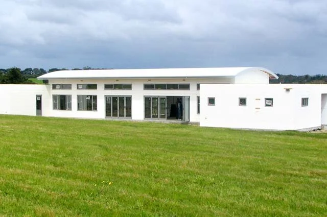 Modern white house with large windows and a curved roof, surrounded by green grass, under a cloudy sky.