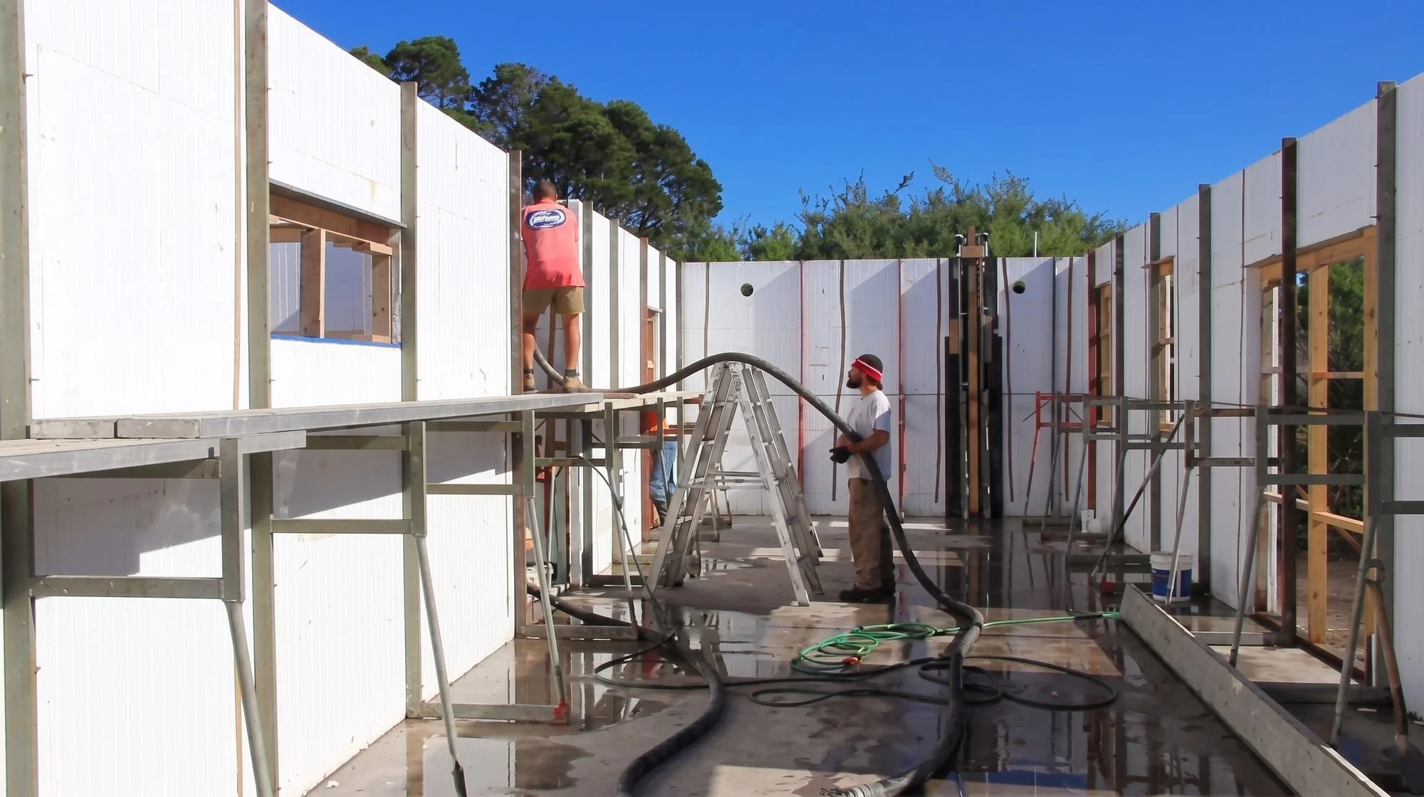Two construction workers are building the interior walls of a house. One worker is standing on stilts, working on the wall, while the other is holding a hose. The structure has wooden framing, and the walls are covered with white insulation panels. The sky is clear and blue, with trees visible in the background.