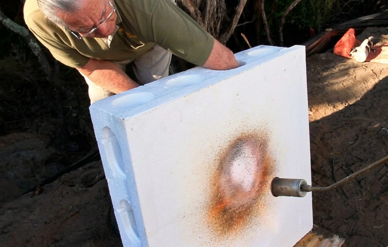 An elderly man is working with a large piece of white foam insulation that is burning or melting from a heat source directed at it. The man is outdoors, wearing glasses and a green shirt.