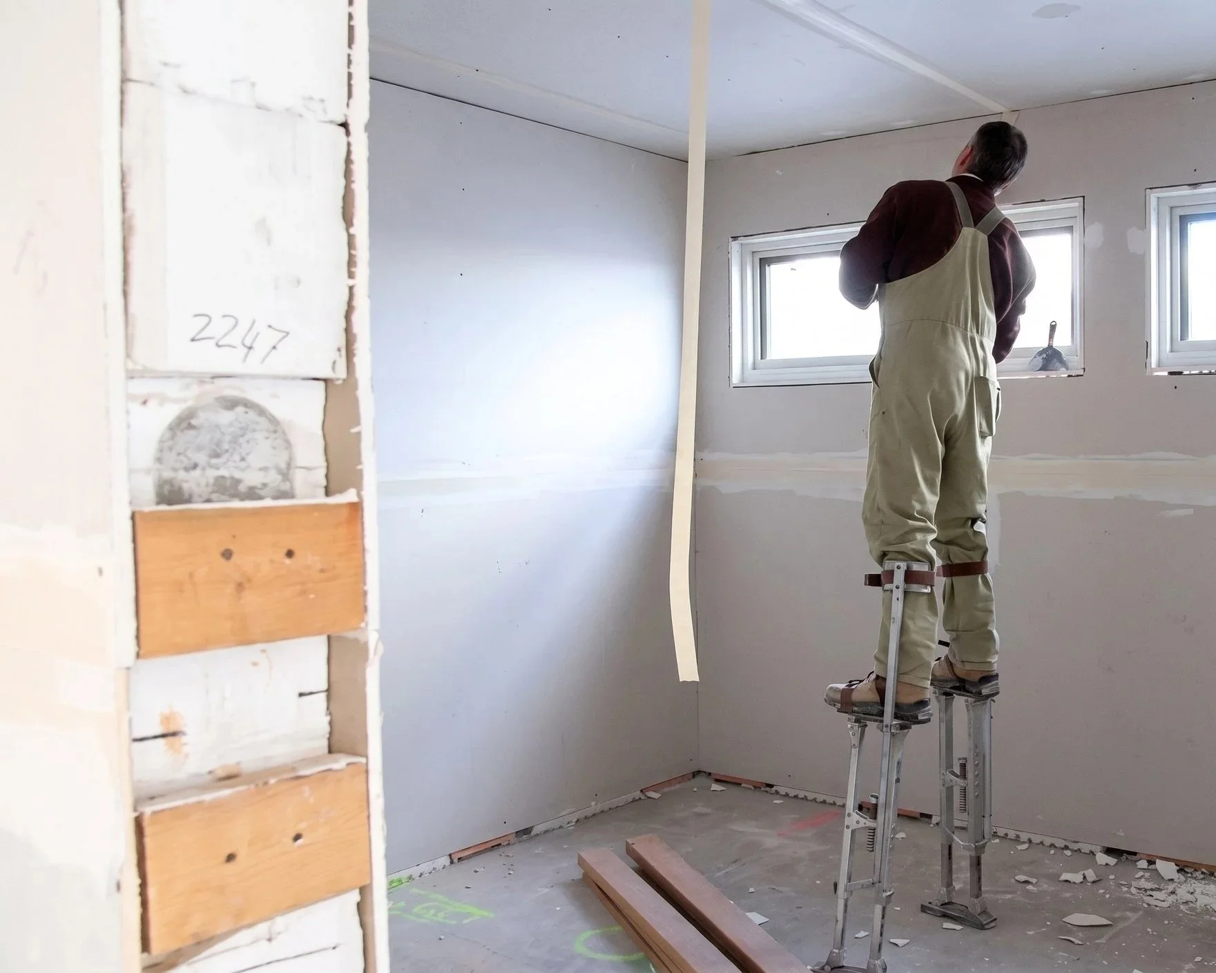 A construction worker stands on a ladder inside a room with unfinished drywall walls and two small windows. He is inspecting the ceiling and wearing safety gear. Building materials and debris are on the floor.