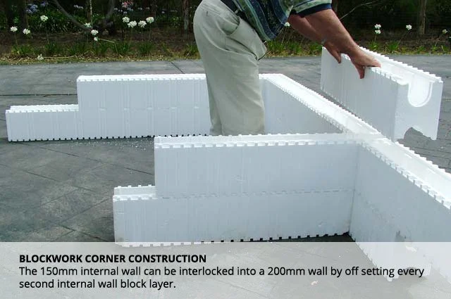 Person assembling white interlocking foam wall blocks outdoors on pavement, with flowering plants in background.