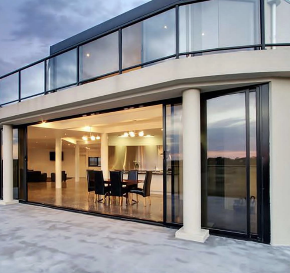 Modern two-story house with large glass sliding doors and an open living area visible inside, featuring a dining table and chairs, overlooking an outdoor patio at sunset.