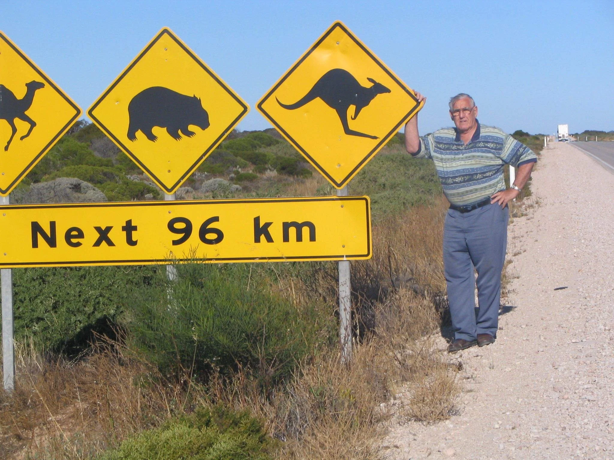 A man standing on the side of a rural road holding a yellow animal crossing warning sign with a kangaroo silhouette. There are other similar signs depicting a camel and a wombat in the background, and a truck approaching on the road.