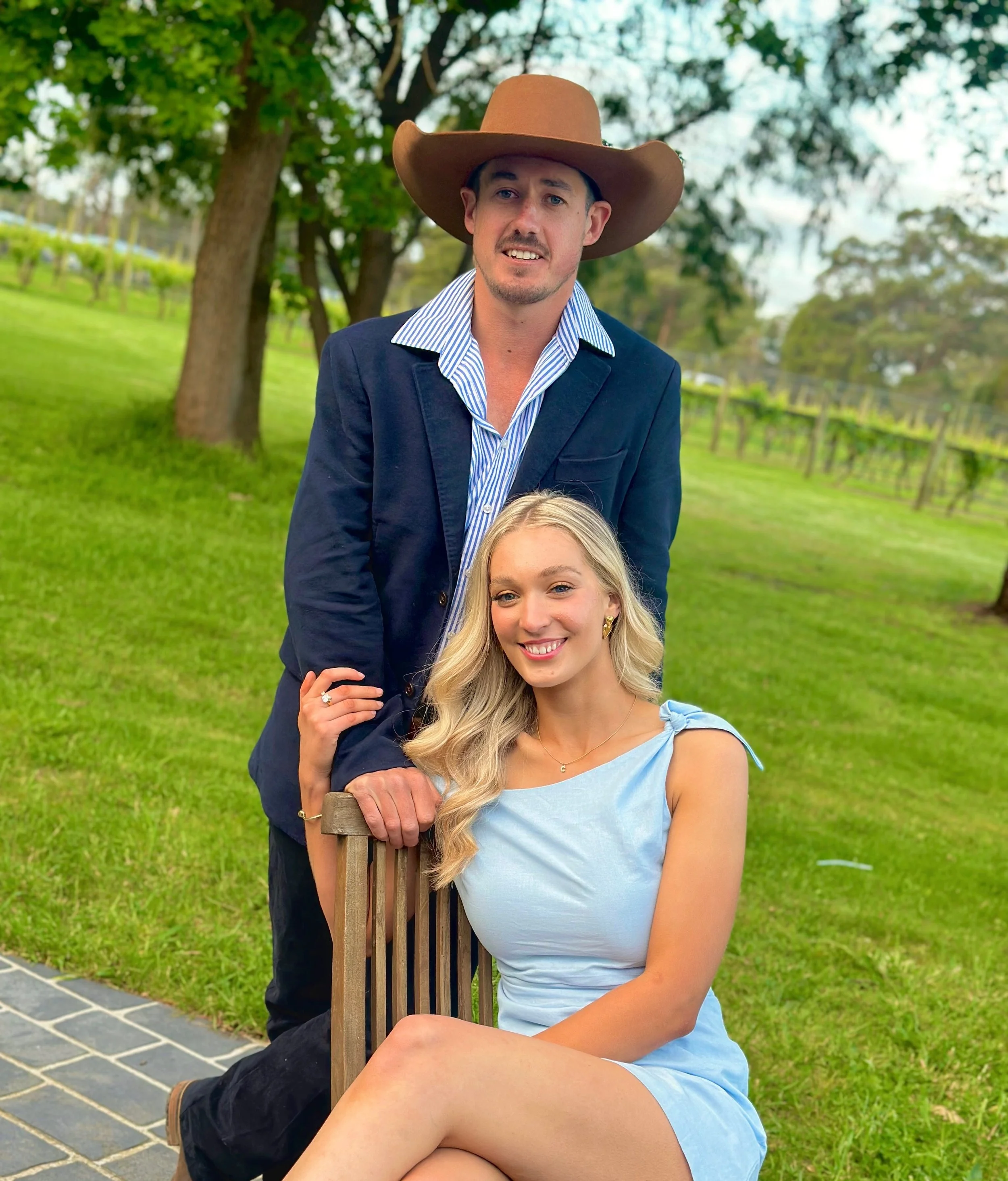 A man wearing a cowboy hat and a woman in a light blue dress sitting outside on a wooden chair with a fence and trees in the background.