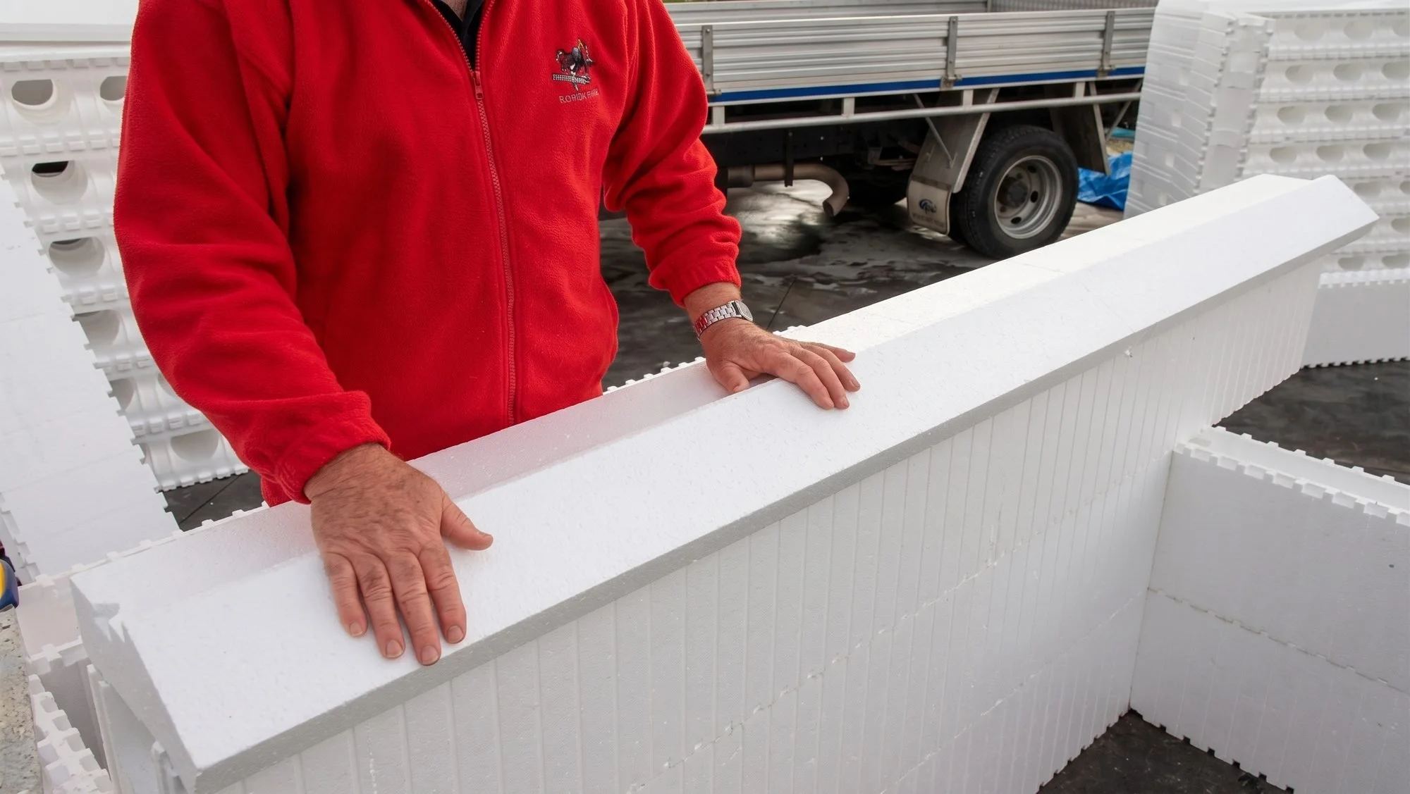 Person in a red jacket holds a white foam block in an outdoor setting with stacked foam blocks and a truck in the background.