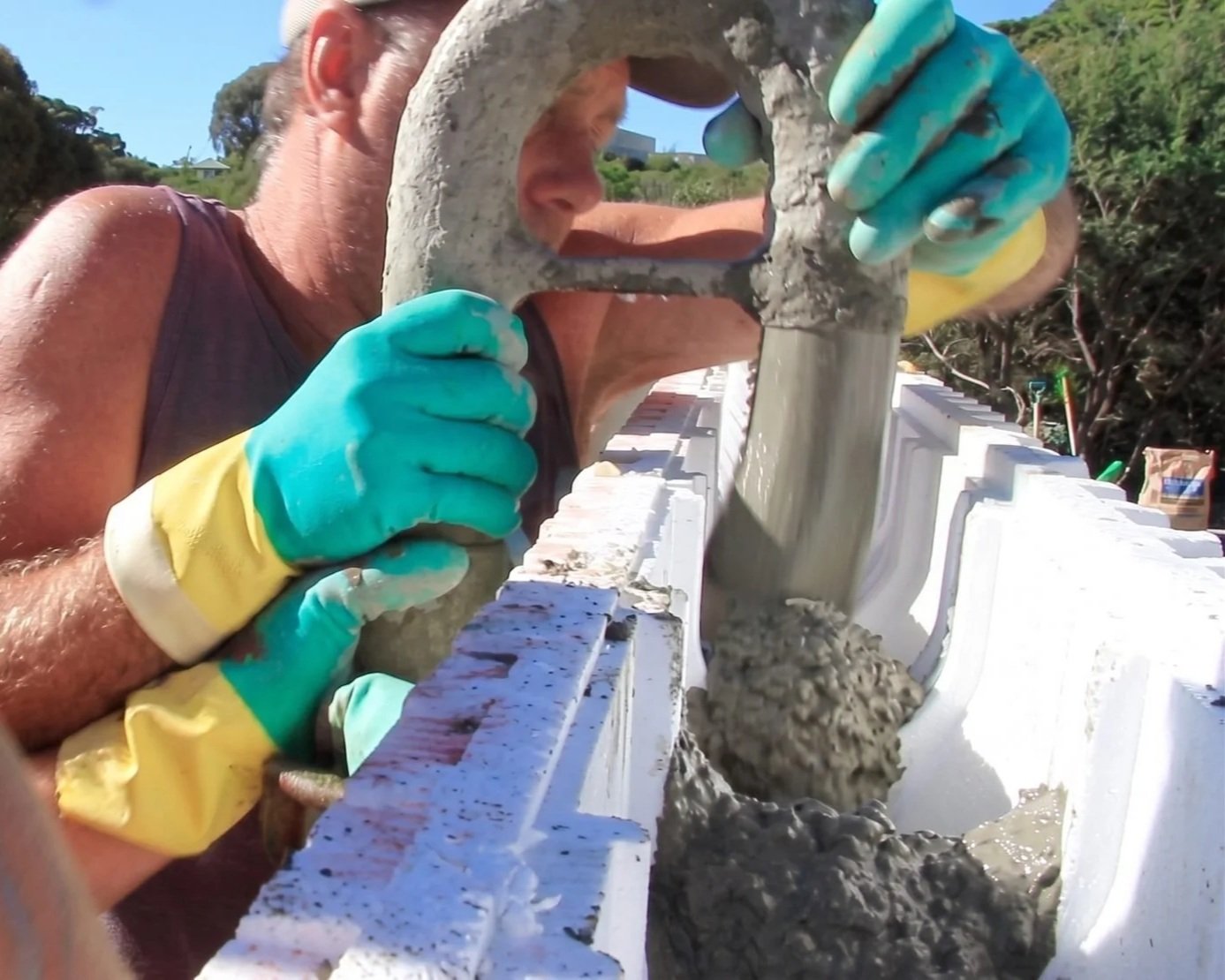 A person wearing yellow and green gloves is working on a construction project, pouring wet concrete into a white concrete mold using a tool, with trees and a clear sky in the background.