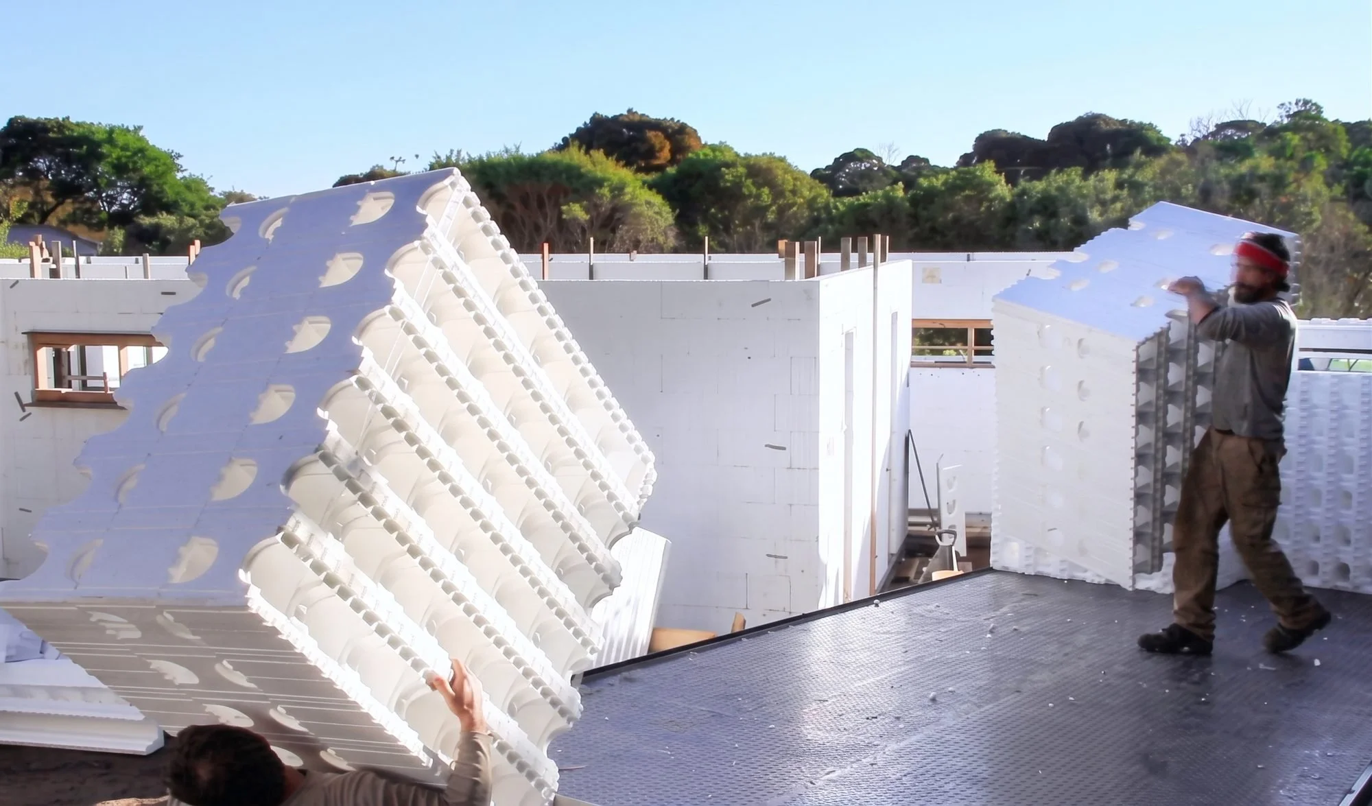 Two workers are installing large white modular insulation panels on the walls of a building under construction during daytime. One worker is holding an insulation panel while another is positioning the panel on the wall. The construction site is outdoors with trees and a clear blue sky in the background.